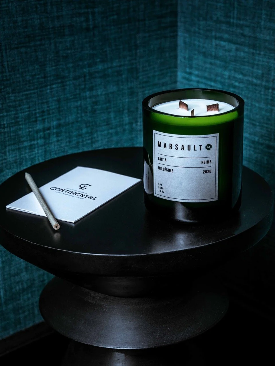 Une bougie verte dans un contenant en verre noir avec une étiquette mentionnant 'Marsault', une assiette noire ronde, un carnet blanc avec un stylo en métal, tous placés sur une petite table noire. L'arrière-plan est une surface rembourrée de tissu b
