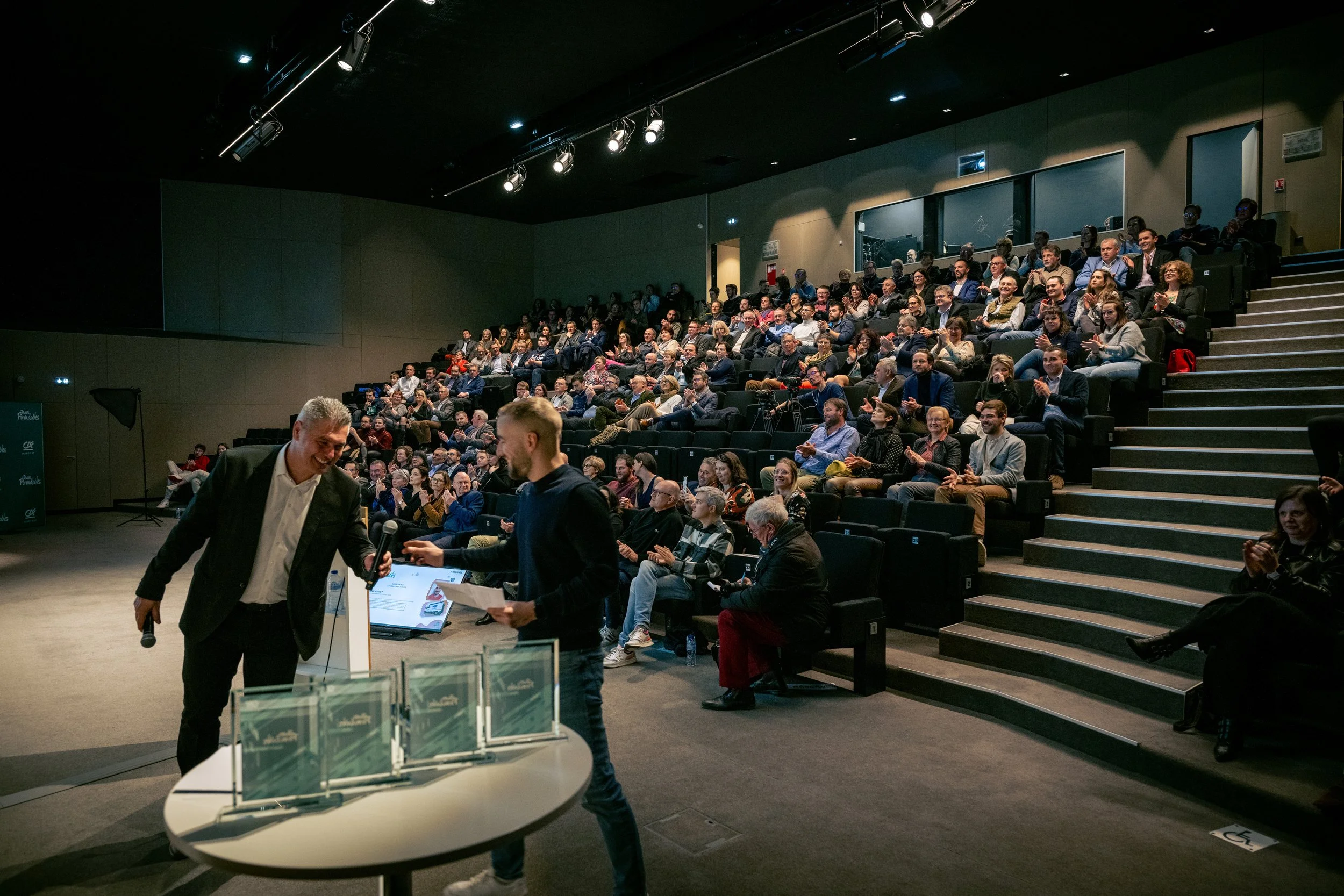 Une salle de conférence ou d'événement remplie de personnes assises en gradins, avec deux hommes au premier plan échangeant un objet, probablement un prix ou une récompense, dans un contexte professionnel ou célébration.