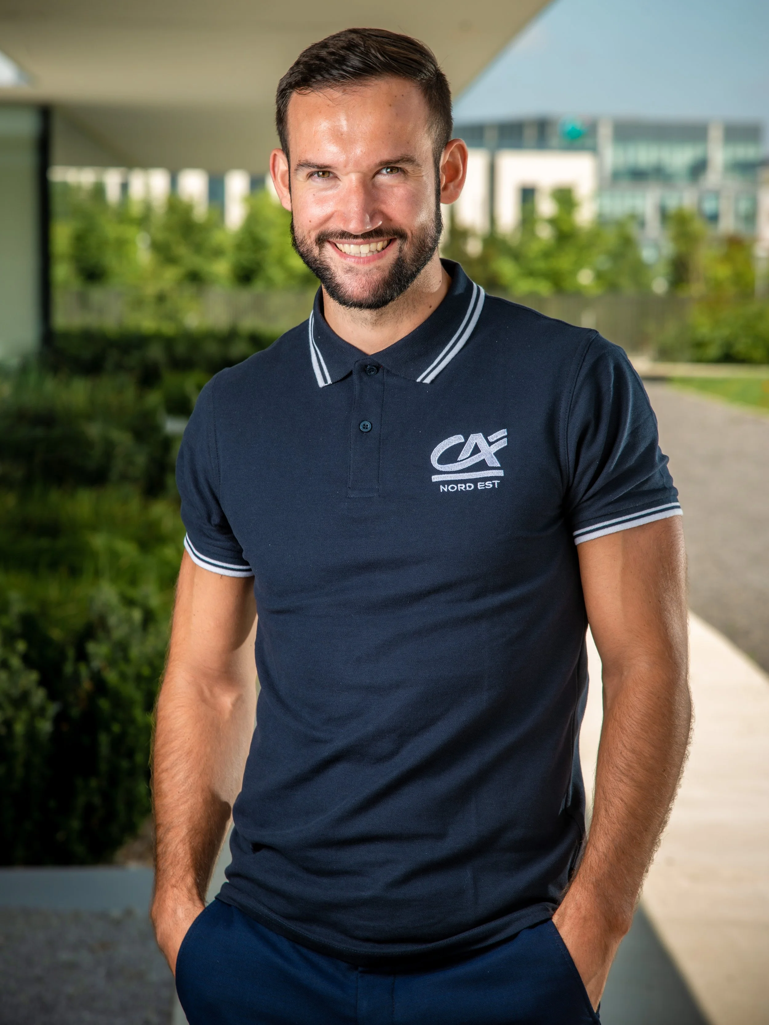 Jeune homme souriant portant un polo noir avec le logo de Crédit Agricole Nord Est, en extérieur urbain. Village by CA Bezannes. Photo prise par Théo WALLYN, TheWallPhoto.