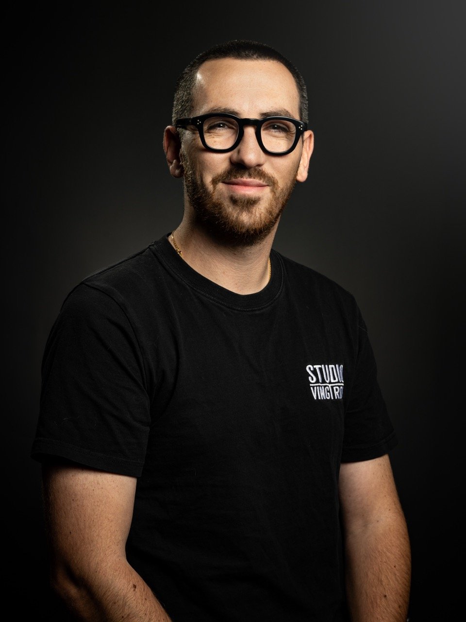 Thomas Scirpo souriant portant des lunettes noires et un t-shirt noir avec le texte "STUDIO VINGT-TROIS" sur la poitrine, posé contre un fond noir. Photo prise par Théo WALLYN, TheWallPhoto.