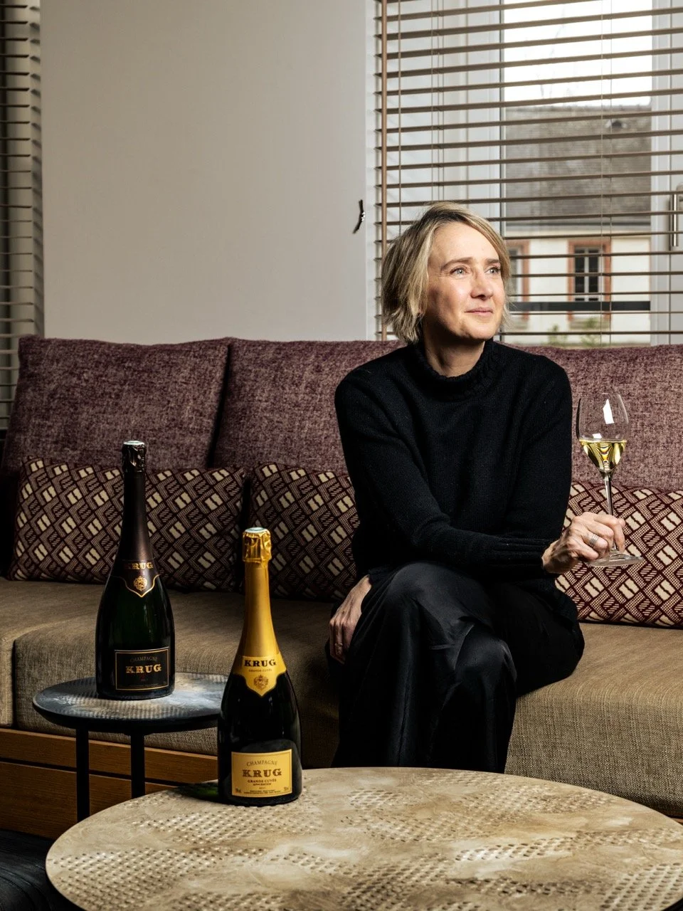 La cheffe de cave Julie Cavil du champagne Krug  assise sur un canapé avec deux bouteilles de champagne Krug et un verre de champagne dans les mains, à l'intérieur d'une pièce avec volets en bois et une fenêtre.