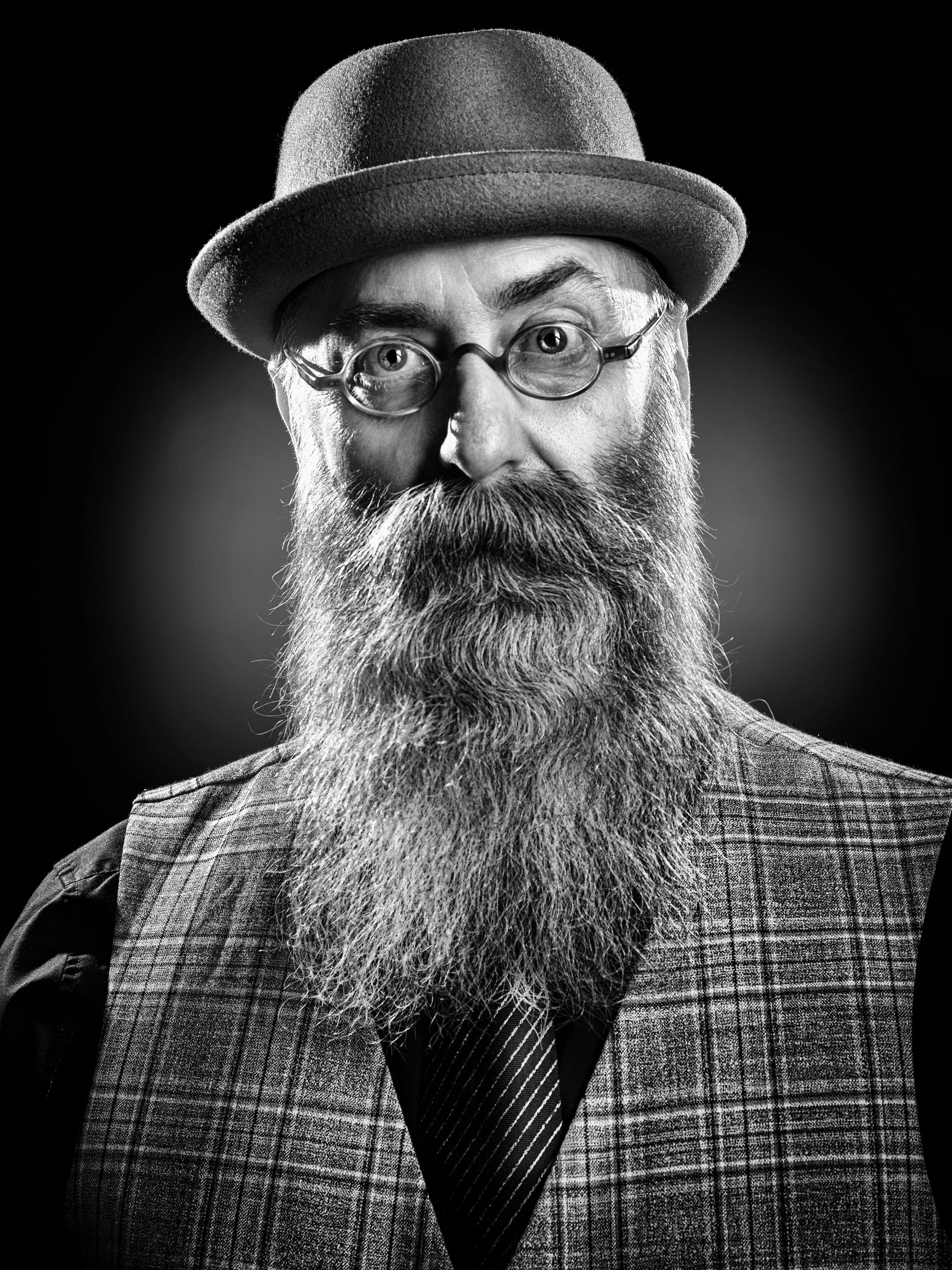 Portrait noir et blanc d'un homme avec une grosse barbe, portant des lunettes rondes, un chapeau et un costume à carreaux.