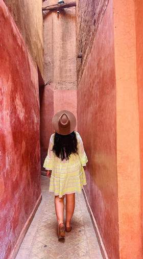 LGBT Woman walking through a narrow alley with pink and red walls, wearing a yellow striped dress and a wide-brimmed hat, seen from behind.