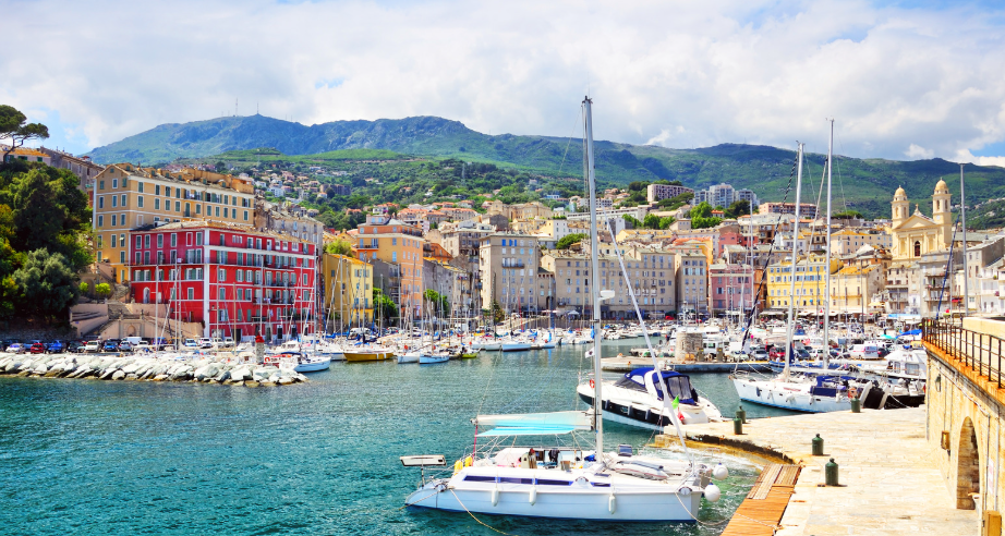 Harbor with sailboats docked and colorful buildings on a hillside, with mountains in the background.
