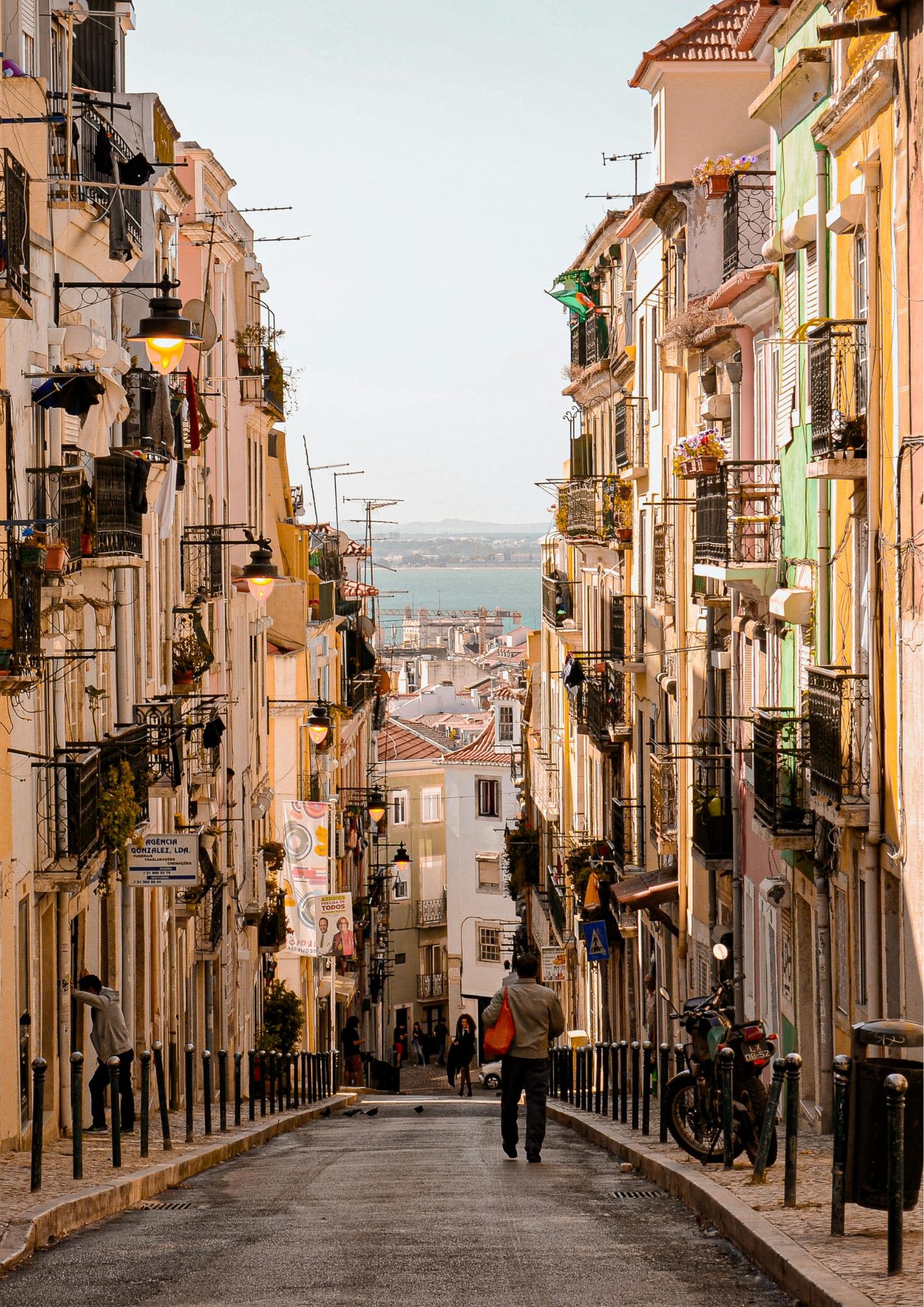 A narrow street in a European city with tall pastel-colored buildings, balconies with laundry and plants, a view of the water and distant land in the background, and a man walking down the street.
