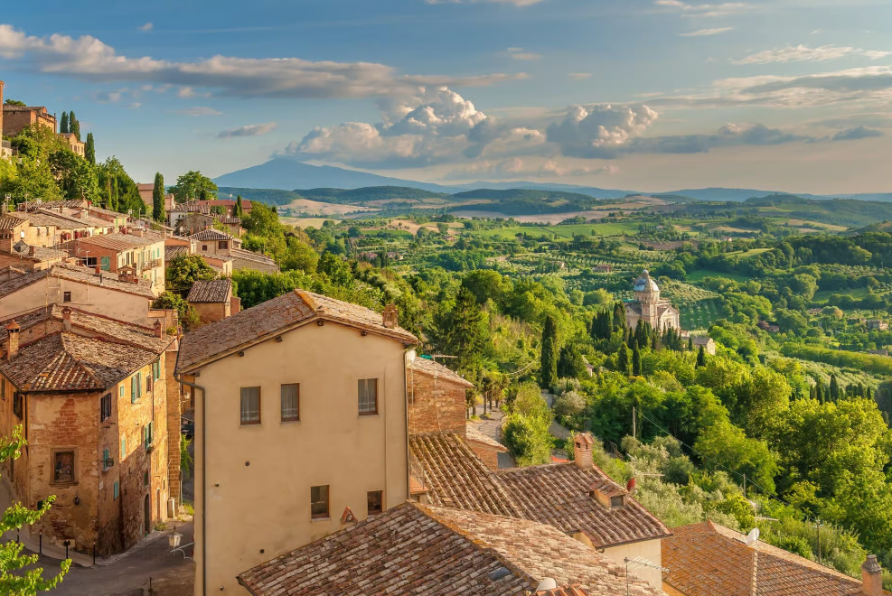 Scenic view of a small Italian hillside town with old buildings, lush greenery, and rolling hills in the background under a partly cloudy sky.