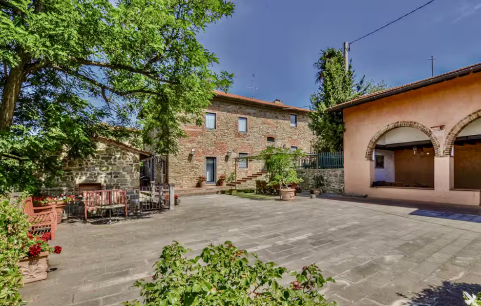 A spacious courtyard surrounded by stone and stucco buildings with potted plants and trees, benches, and a small outdoor fireplace.