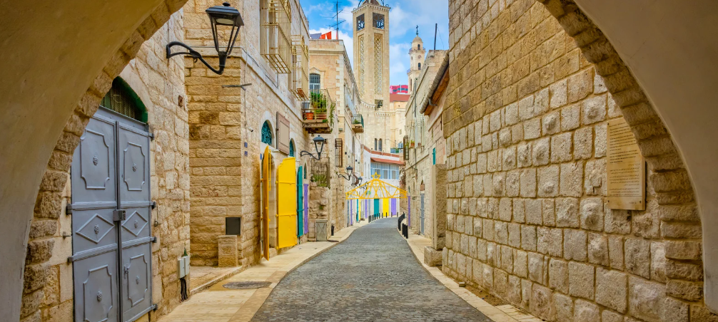 Narrow cobblestone street lined with beige stone buildings, colorful doors, and black wrought iron lamps, leading towards a tower with flags, in a sunny Mediterranean setting.