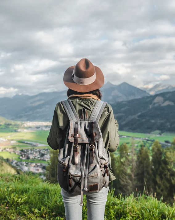 A LGBT woman standing on a grassy hill overlooking a valley with a town and mountains in the background, wearing a brown hat, green jacket, and carrying a backpack.
