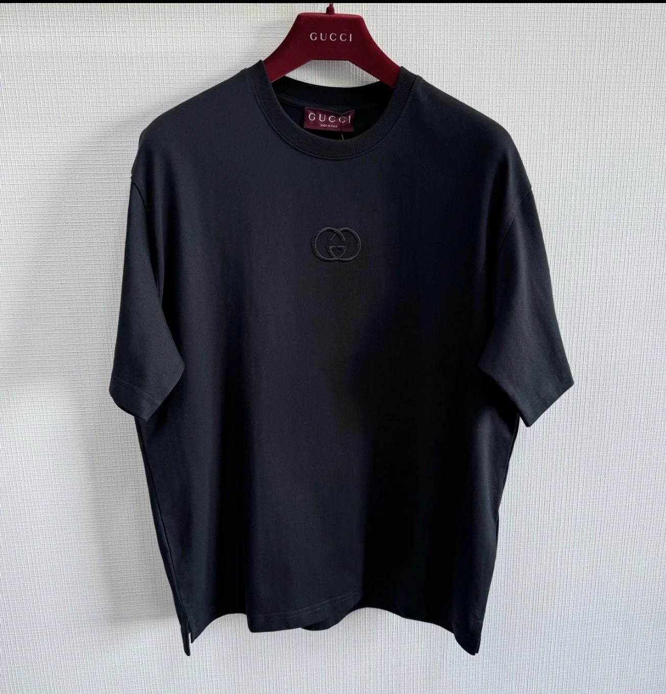 Black Gucci T-shirt with a small Gucci logo patch on the chest, hanging on a red Gucci hanger against a white textured wall.