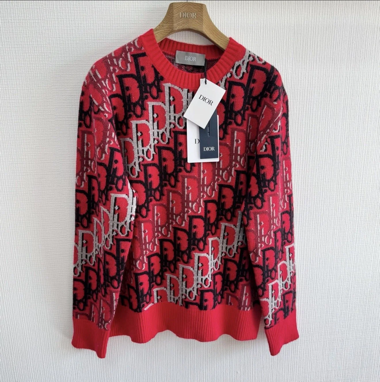 Red sweater with black, gray, and white graphic pattern hanging on a Dior-branded wooden hanger against a plain white wall.