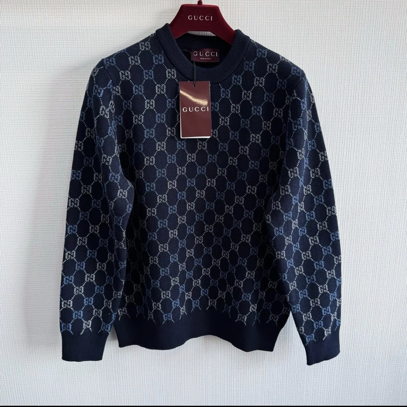A navy blue Gucci sweater with the brand's signature interlocking G pattern in light blue and white, hanging on a red Gucci-branded hanger against a plain wall.