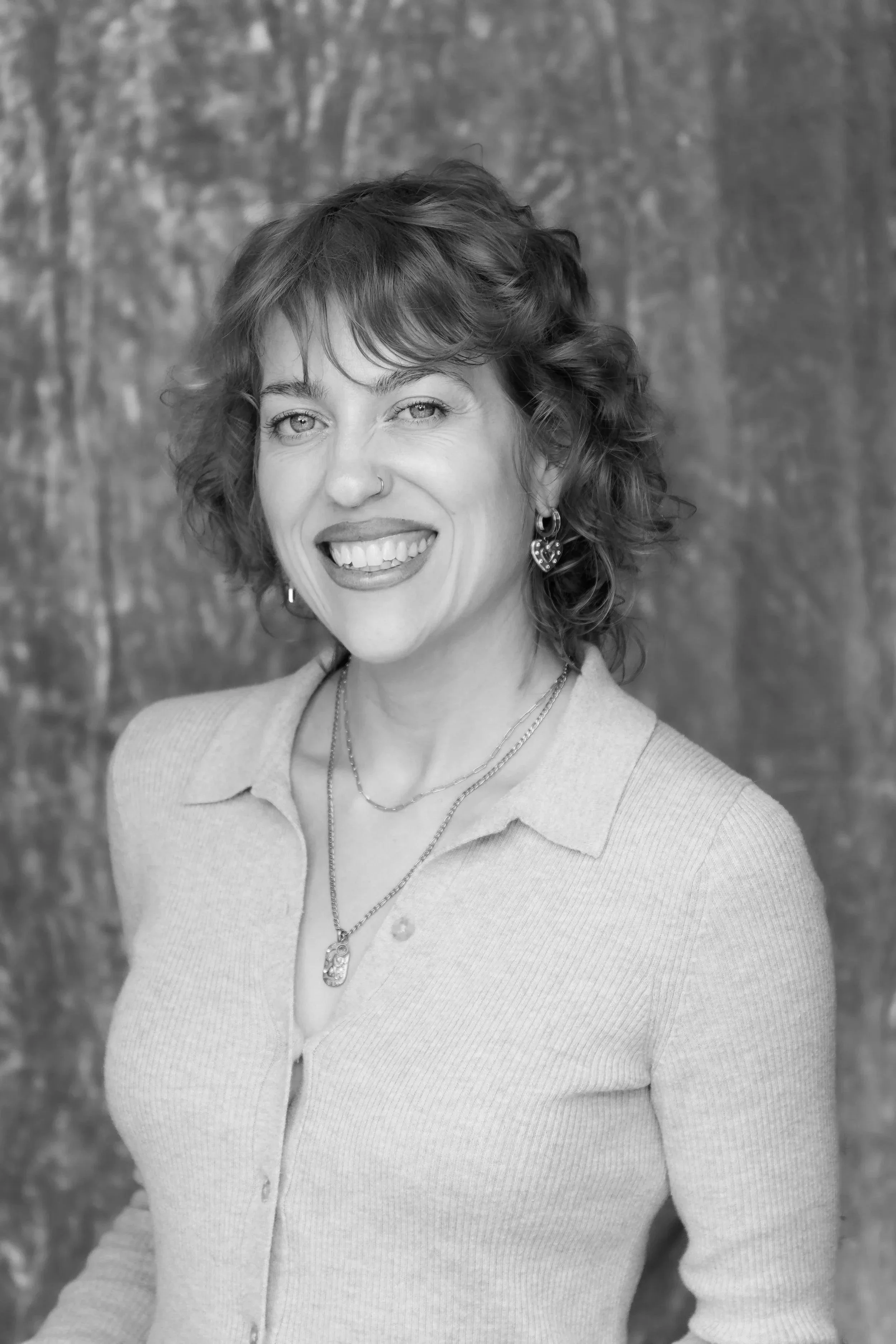 Black and white portrait of a woman with curly hair, smiling, wearing a light-colored button-up shirt and jewelry, standing against a textured background.
