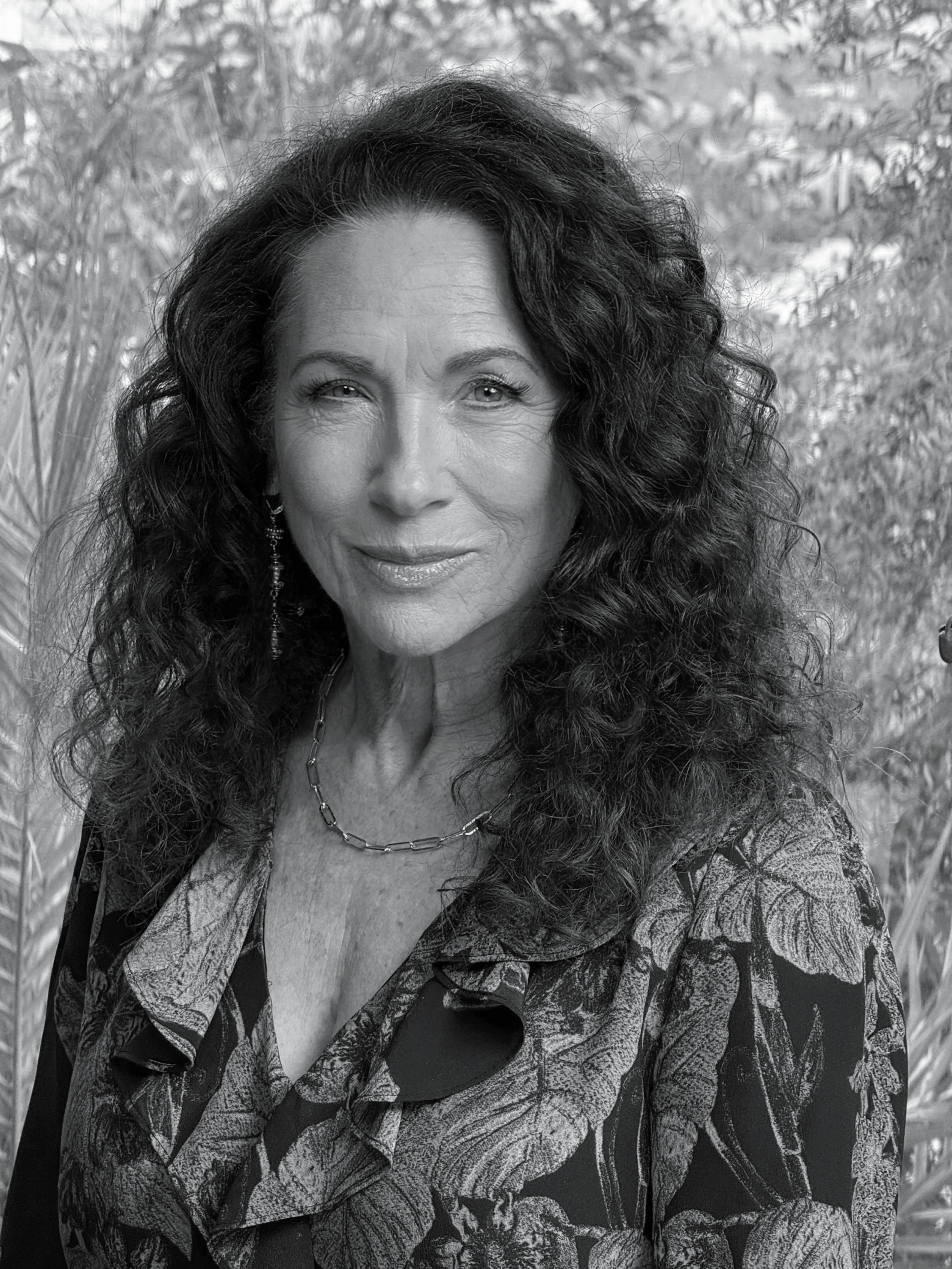 A black-and-white portrait of a woman with long, curly hair, wearing earrings, a necklace, and a patterned blouse, outdoors with plants in the background.
