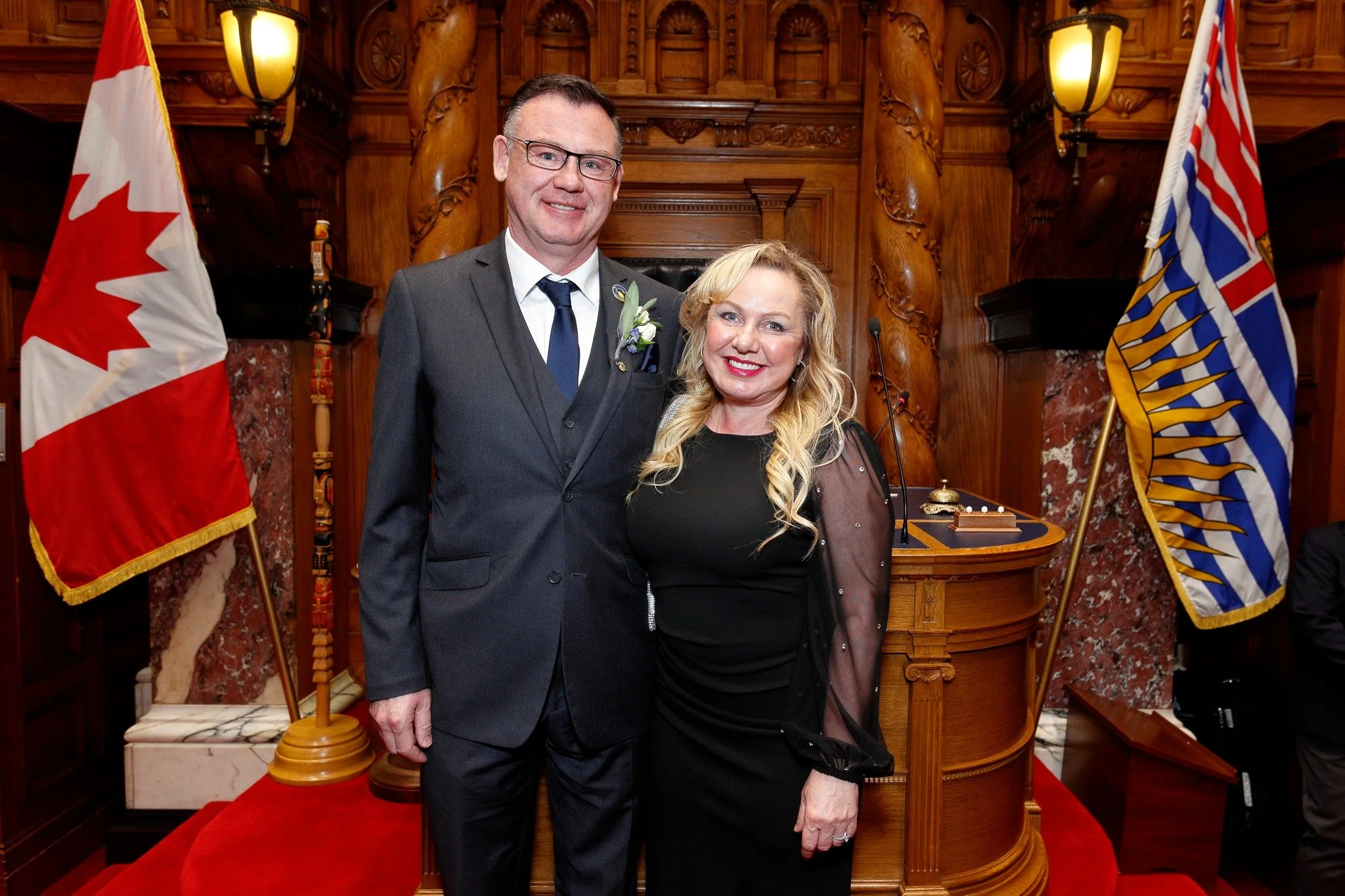 A man in a suit and a woman in a black dress standing together in a formal setting with Canadian and British Columbian flags behind them.