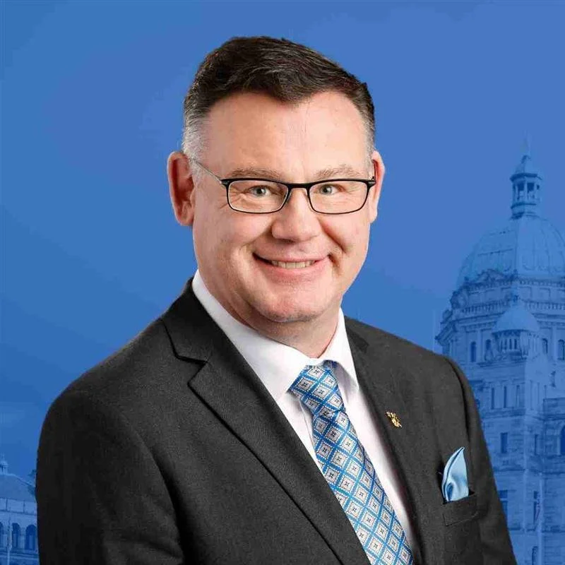 A man wearing glasses, a black suit, white shirt, and blue patterned tie, smiling in front of a blue background with a faint outline of a historic government building.