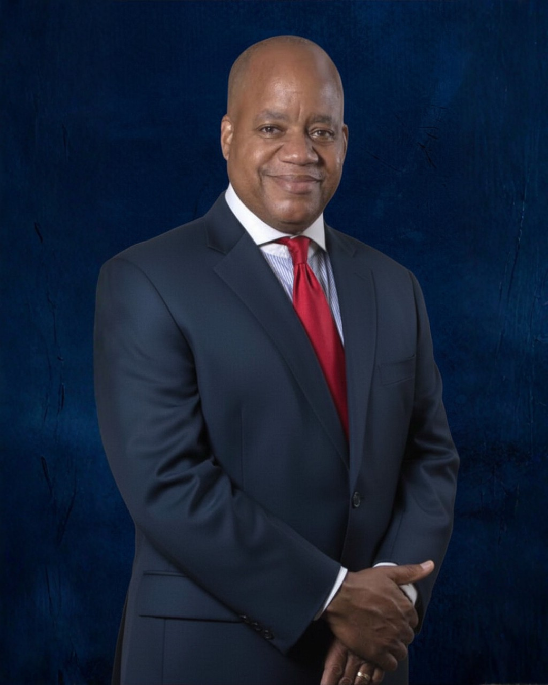 A man in a dark suit, white dress shirt, and red tie standing against a dark blue textured background.