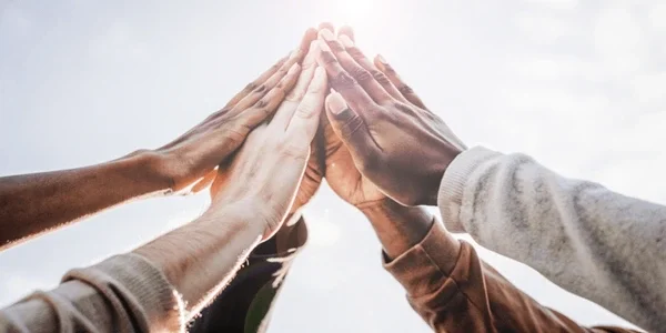 Multiple hands touching together in a high-five or team gesture against a cloudy sky.