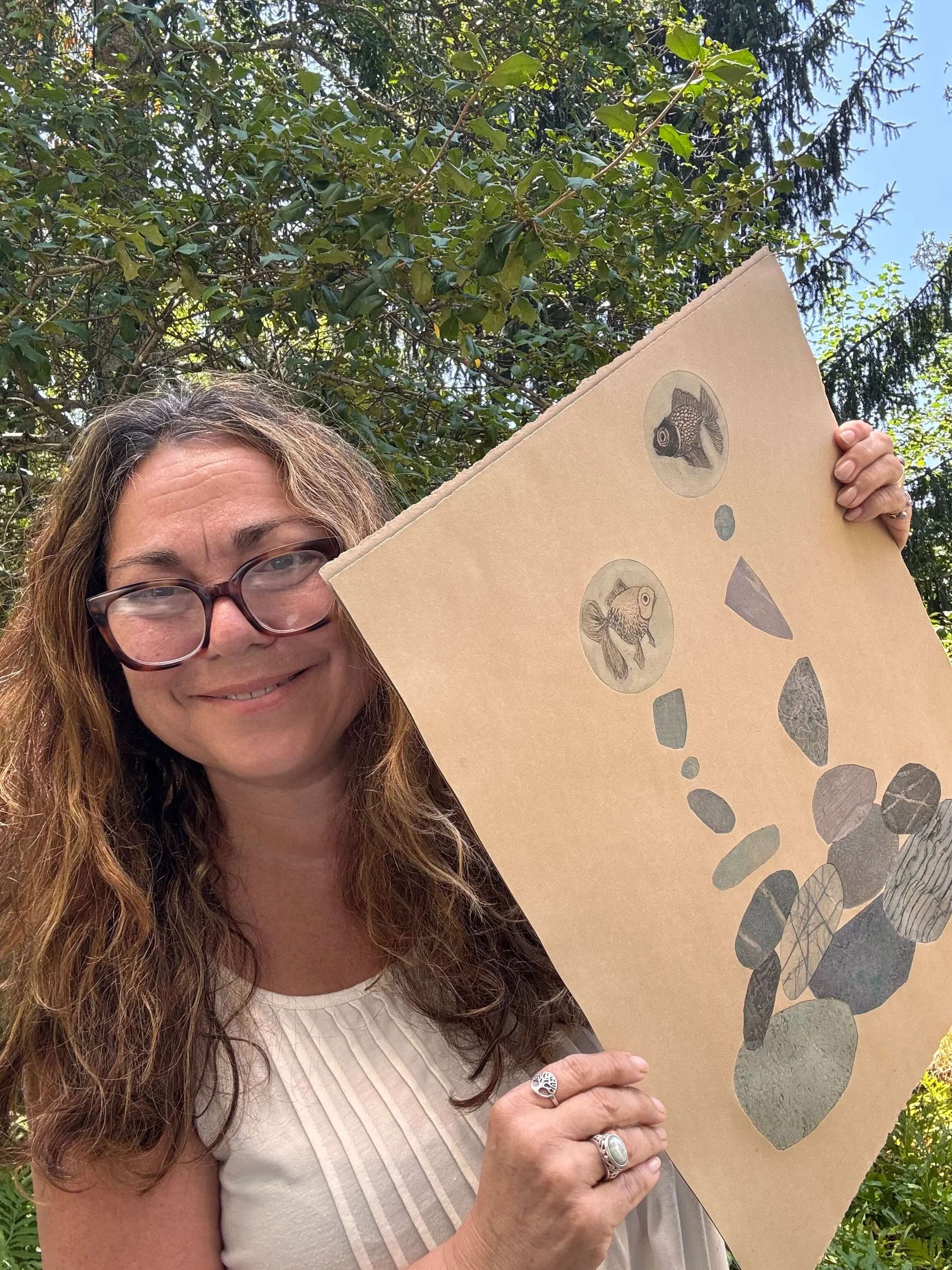 A smiling woman with glasses and long wavy hair holding a piece of tan Stonehenge 100% cotton paper with etching of fish and rocks outdoors.