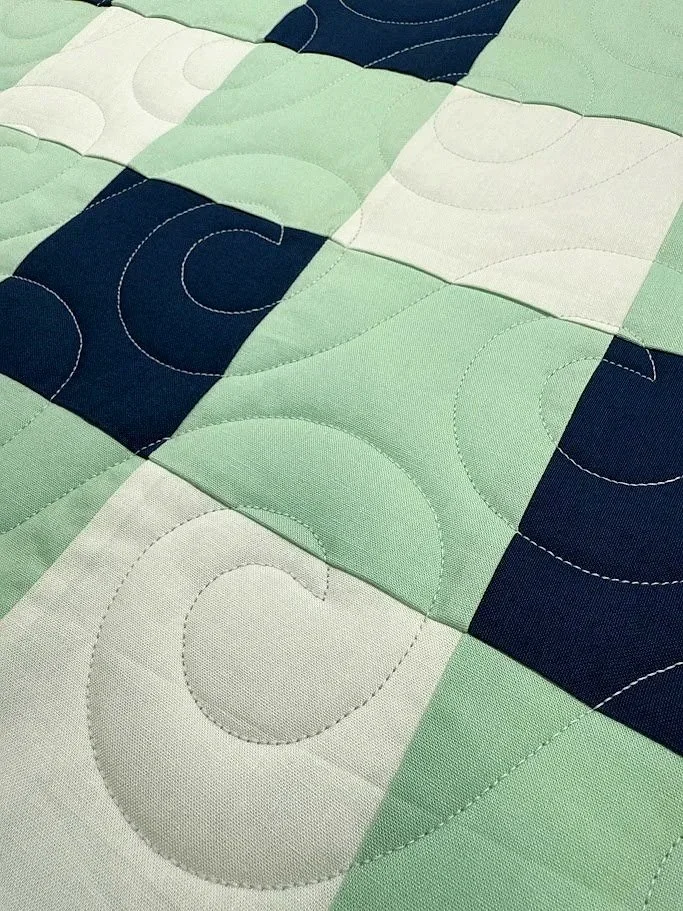 Close-up of a quilt with a pattern of green, navy blue, and white squares, featuring stitched swirling and curved lines.