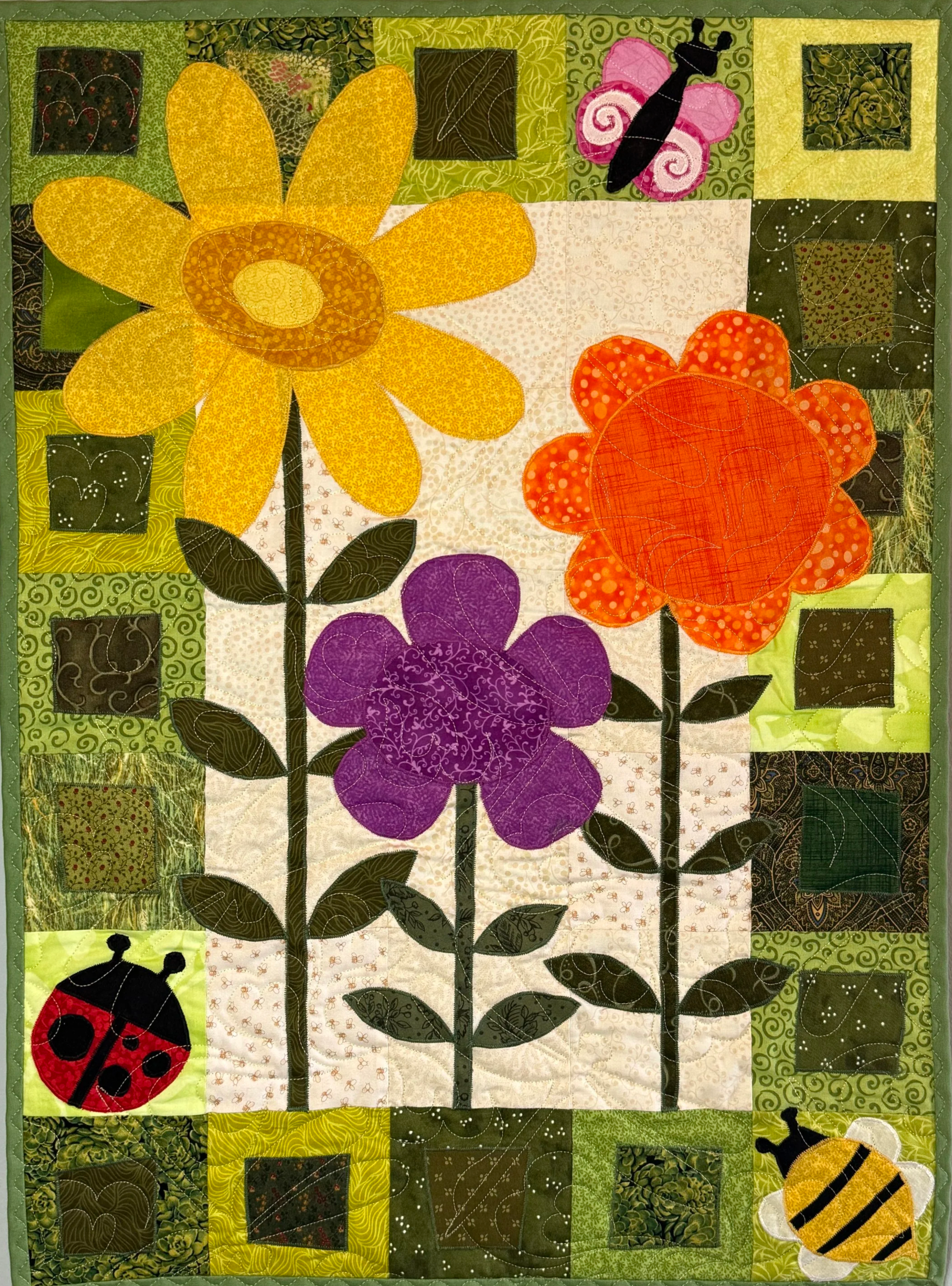 A colorful patchwork quilt with large flowers, ladybugs, and bees, featuring green, yellow, orange, purple, pink, and black quilted fabric pieces.