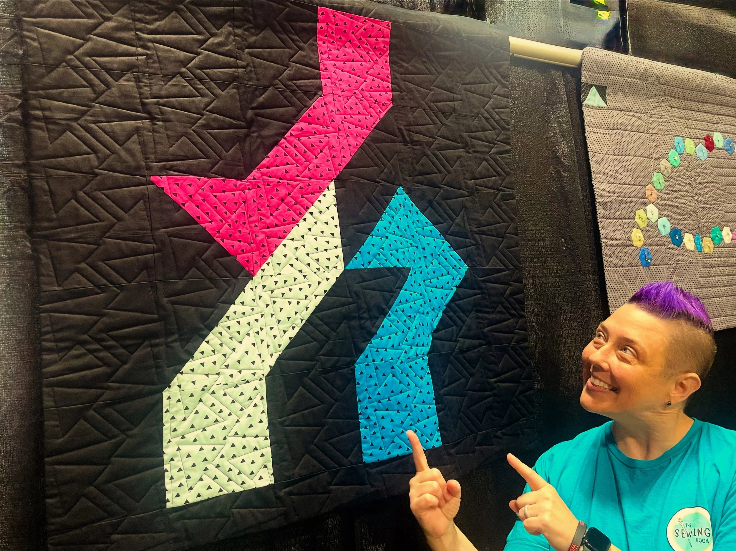 A person with purple hair, glasses, and a bright blue shirt smiling and pointing at a colorful quilt with geometric patterns and triangles in pink, white, blue, and turquoise, with a black background.