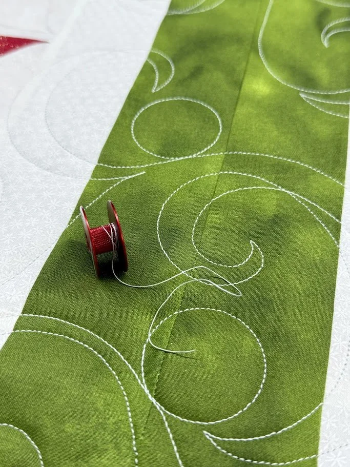 Green fabric with white embroidered circular patterns, sewing needle with red thread resting on it, and additional fabric pieces in white and light gray with similar embroidery.