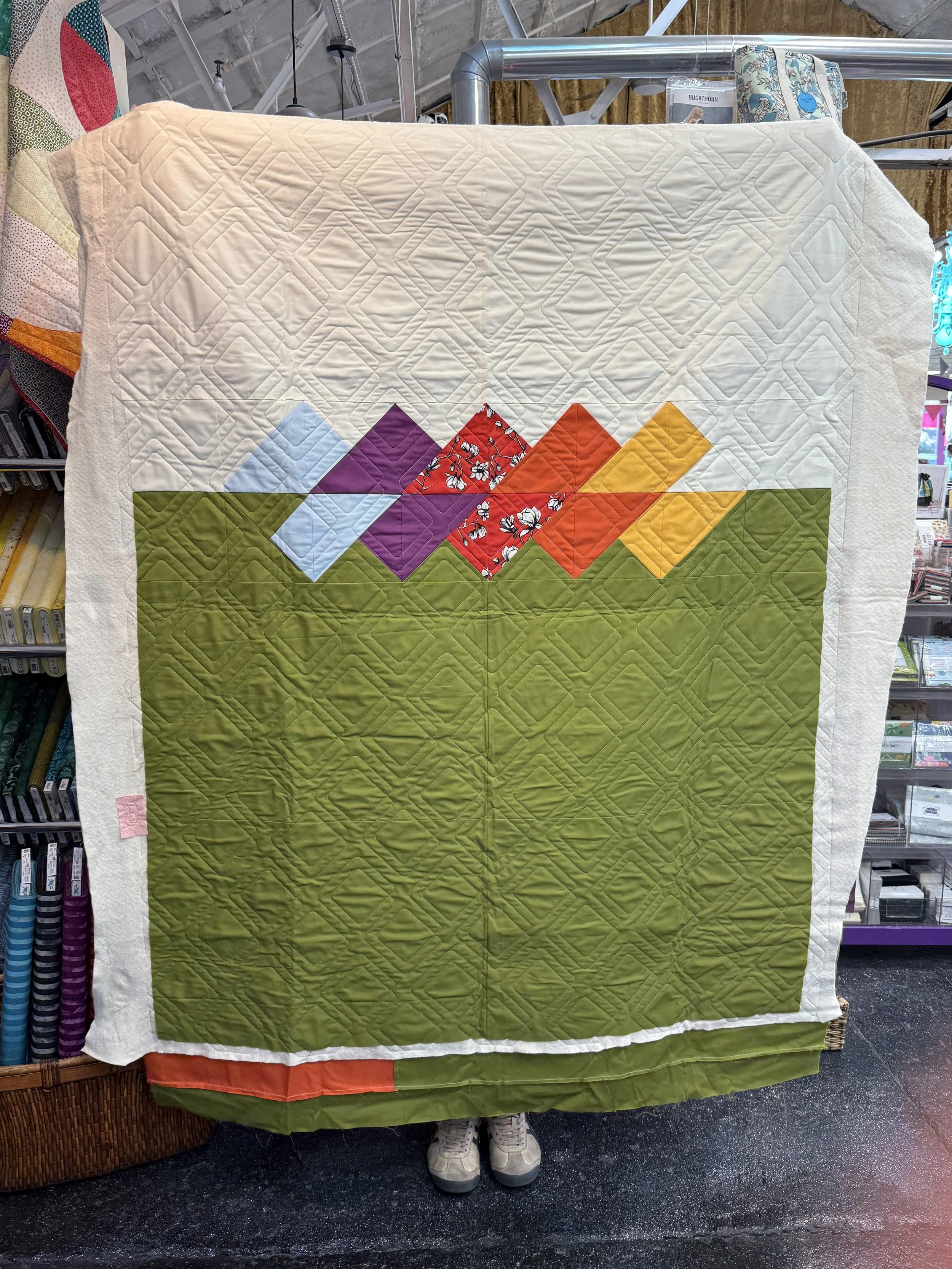 A person holding up a patchwork quilt that covers them from head to toe, showing geometric block patterns in various colors such as purple, red, orange, yellow, green, and blue.