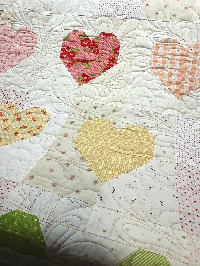 Close-up of a quilt featuring various pieced fabric hearts in different colors and patterns, with intricate quilting stitches and white background fabric.
