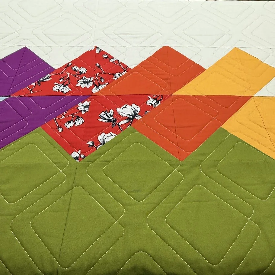 Colorful quilted placemats in purple, red with white flowers, orange, yellow, and green, arranged on a white surface.