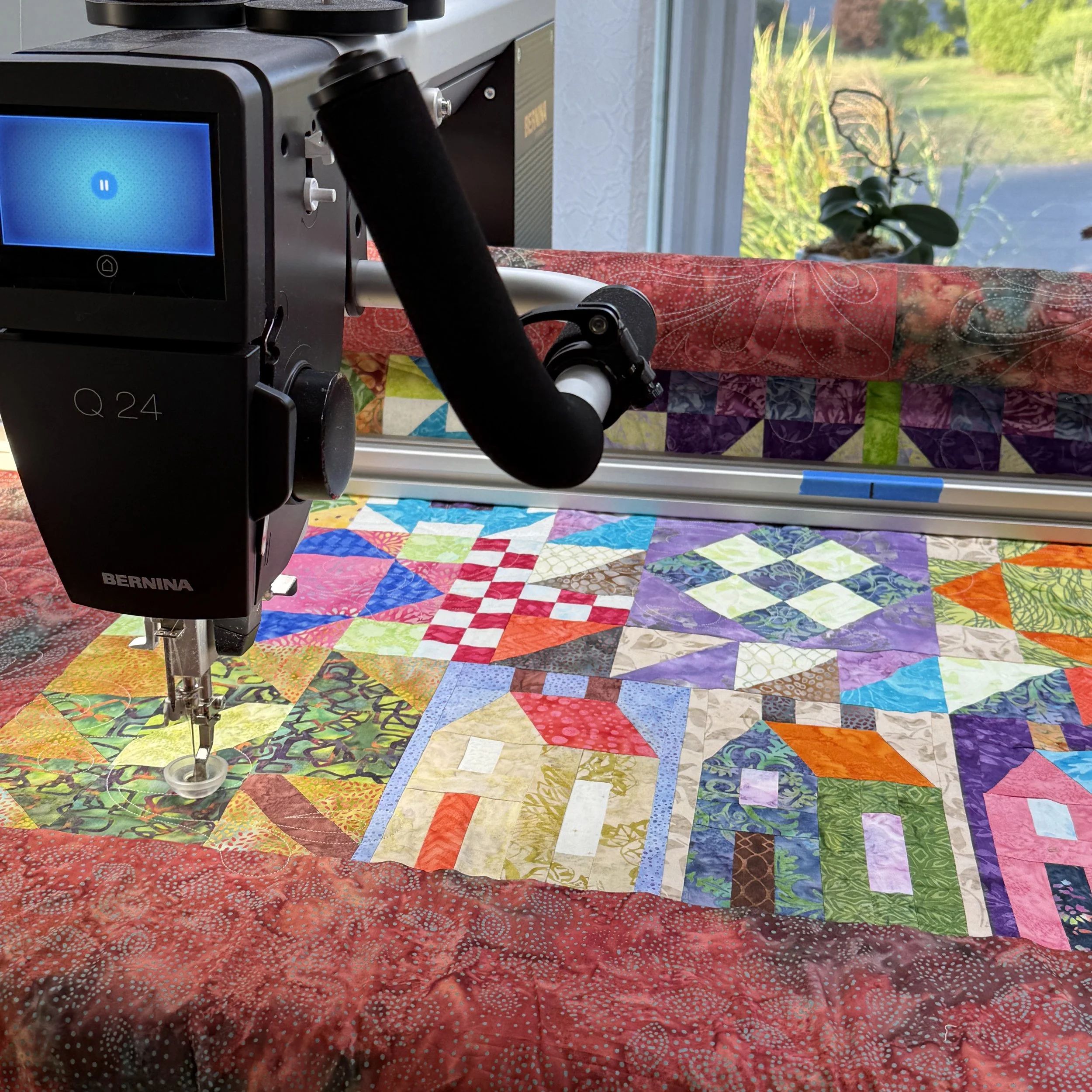 Close-up of a quilting sewing machine and colorful patchwork quilt with various patterns and colors, near a window with outside greenery.