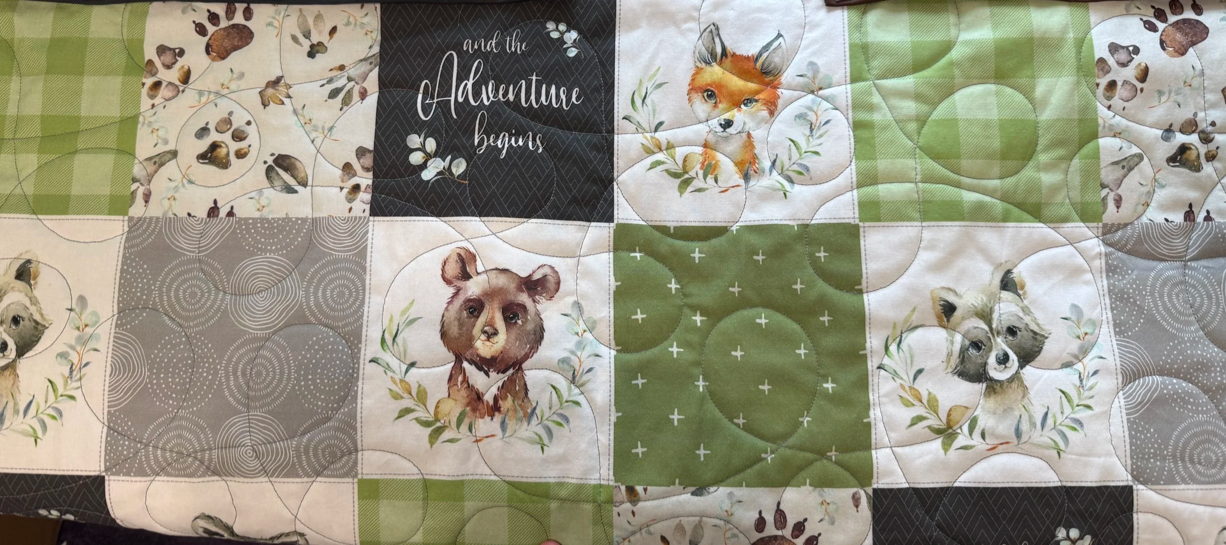 Patterned quilt with illustrations of various dog and fox faces, and a black square with white cursive text reading 'and the Adventure begins'.