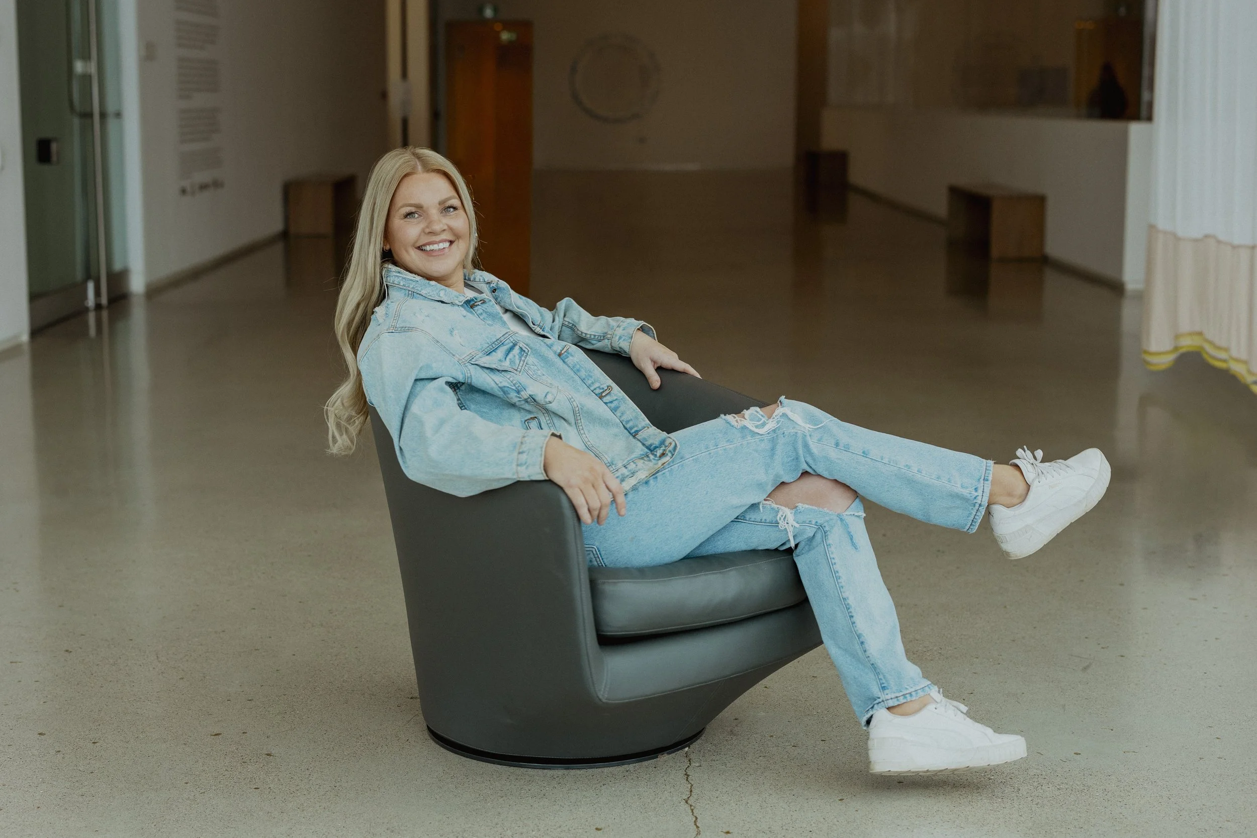 A smiling woman with blonde hair, dressed in a blue denim jacket and ripped jeans, sitting casually in a modern, black lounge chair in an empty indoor space.