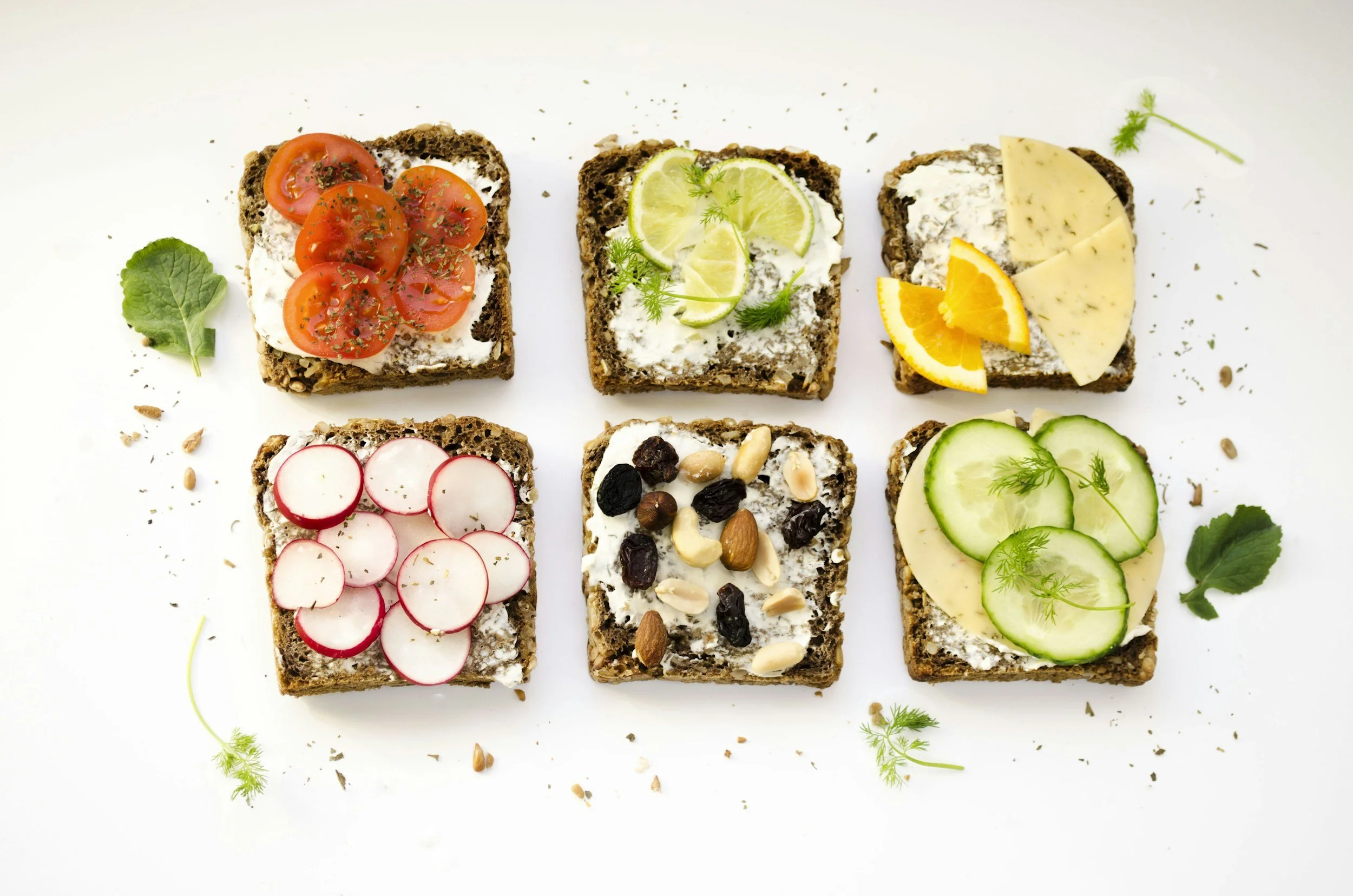 Six whole grain bread slices topped with various spreads and garnishes, arranged in two rows of three, with herbs, seeds, and small leaves scattered around on a white background.