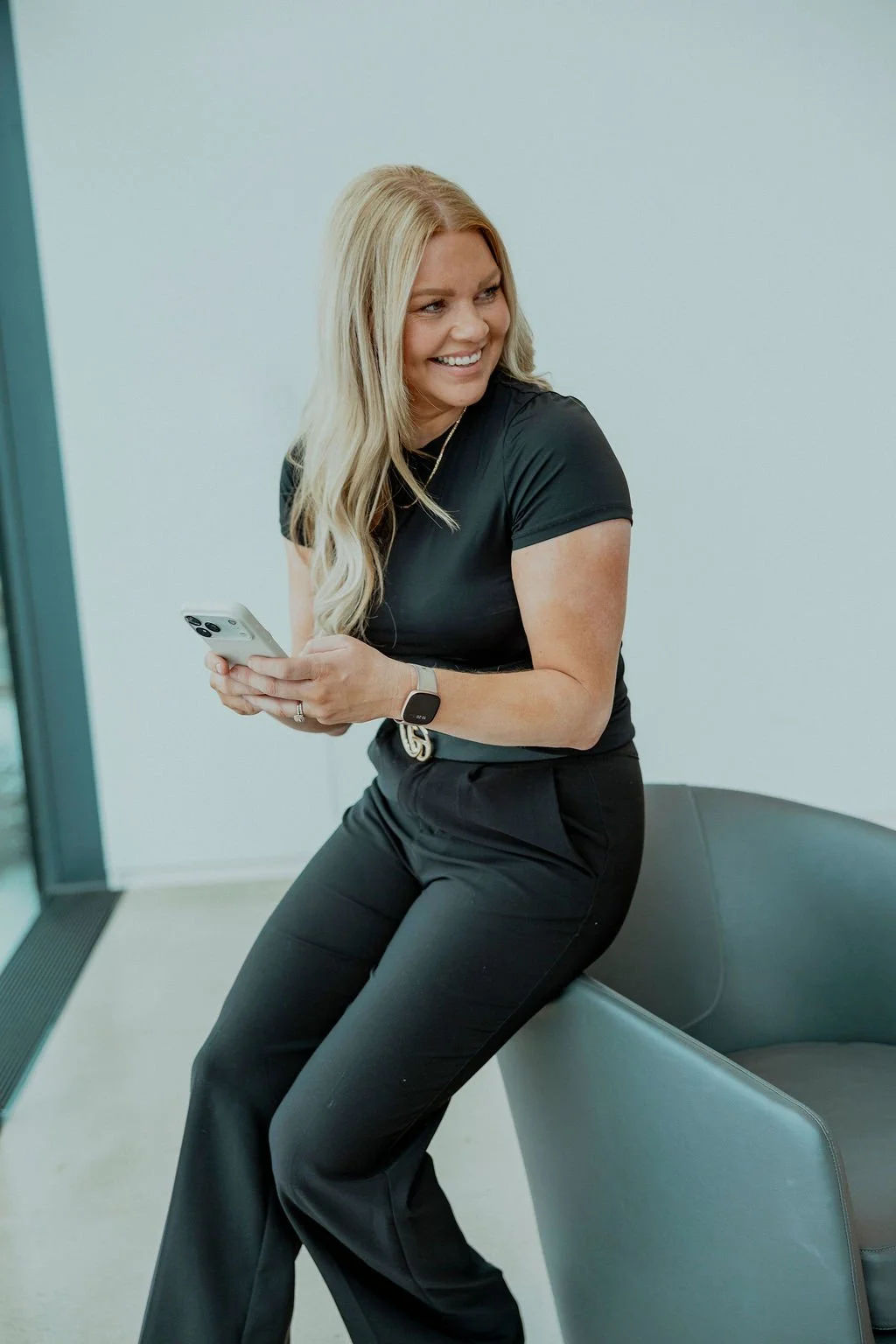 A woman with long blonde hair smiling while holding a smartphone, sitting on a gray chair in a modern office.