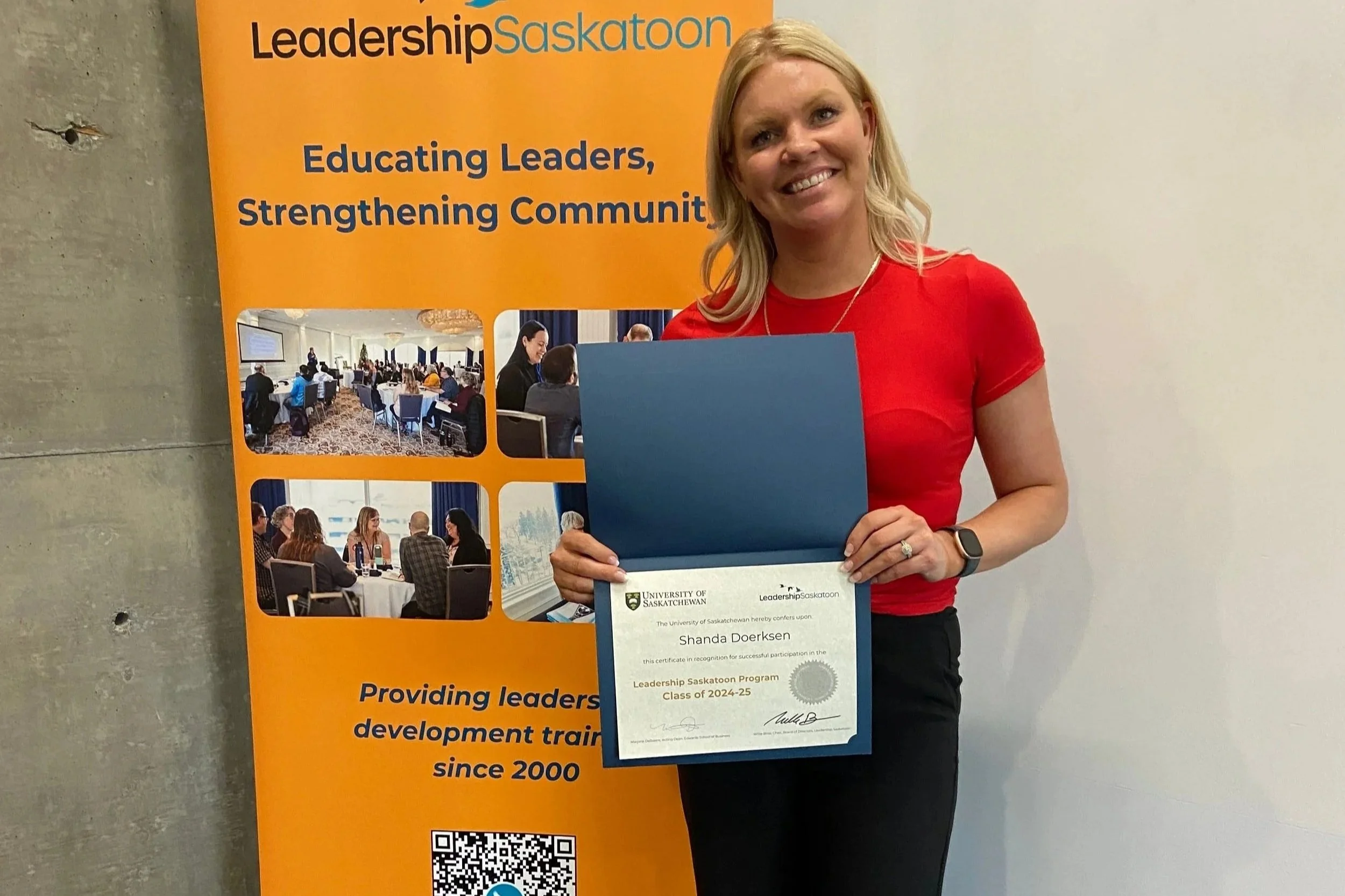 A woman with blonde hair smiling and holding a certificate in front of an orange banner that reads 'Leadership Saskatchewan' and 'Educating Leaders, Strengthening Communities'. The certificate is from the University of Saskatchewan recognizing her participation in the Leadership Saskatchewan Program, Class of 2024-25.