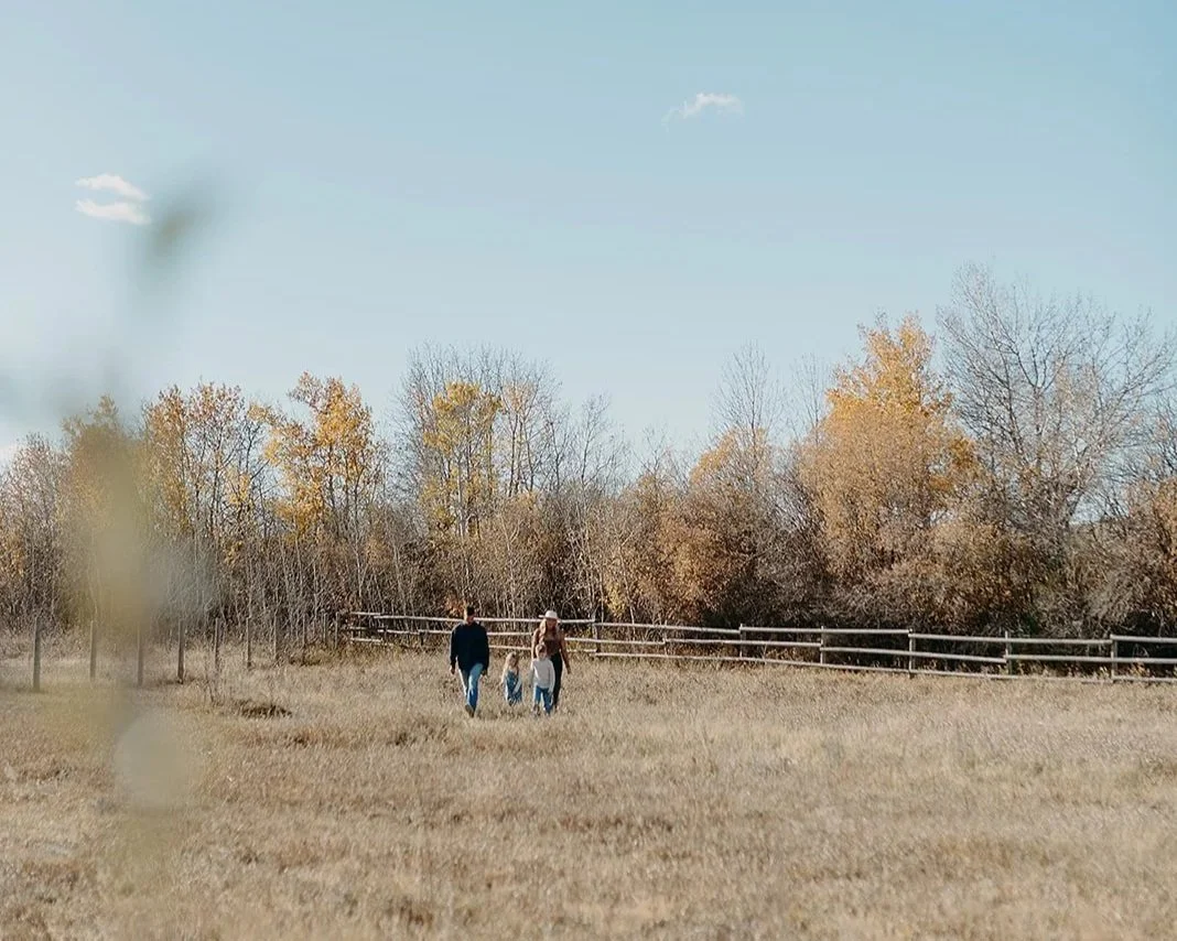 Family walking together in a grassy field during fall, with trees showing autumn colors and a wooden fence in the background.