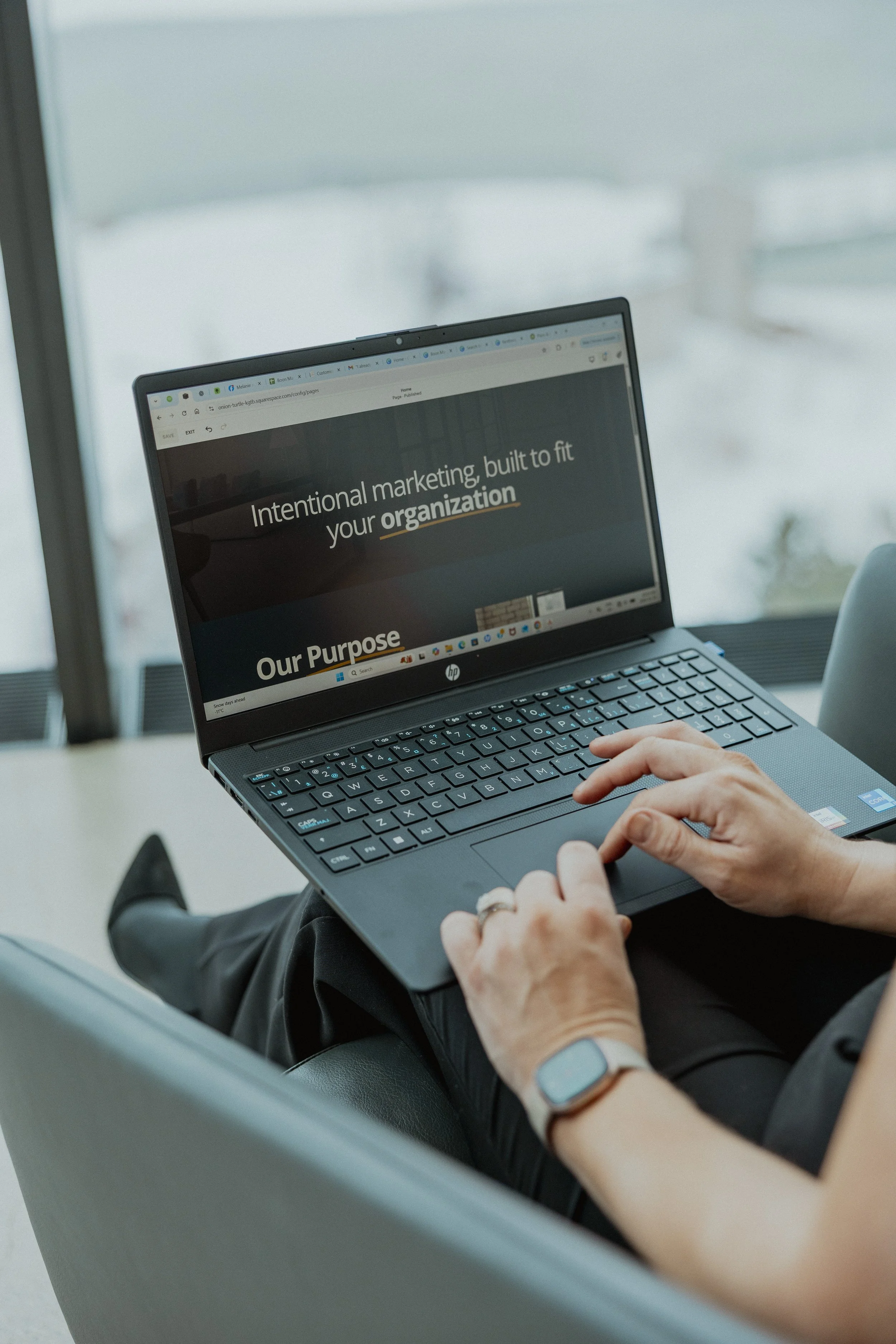 Person sitting on a gray chair using a black laptop with a website open that reads 'Intentional marketing, built to fit your organization' on a large windowed building.