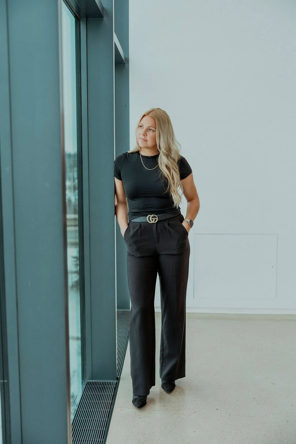 A woman with long blonde hair in a black outfit standing near a large window, looking thoughtfully outside, in a modern indoor setting.