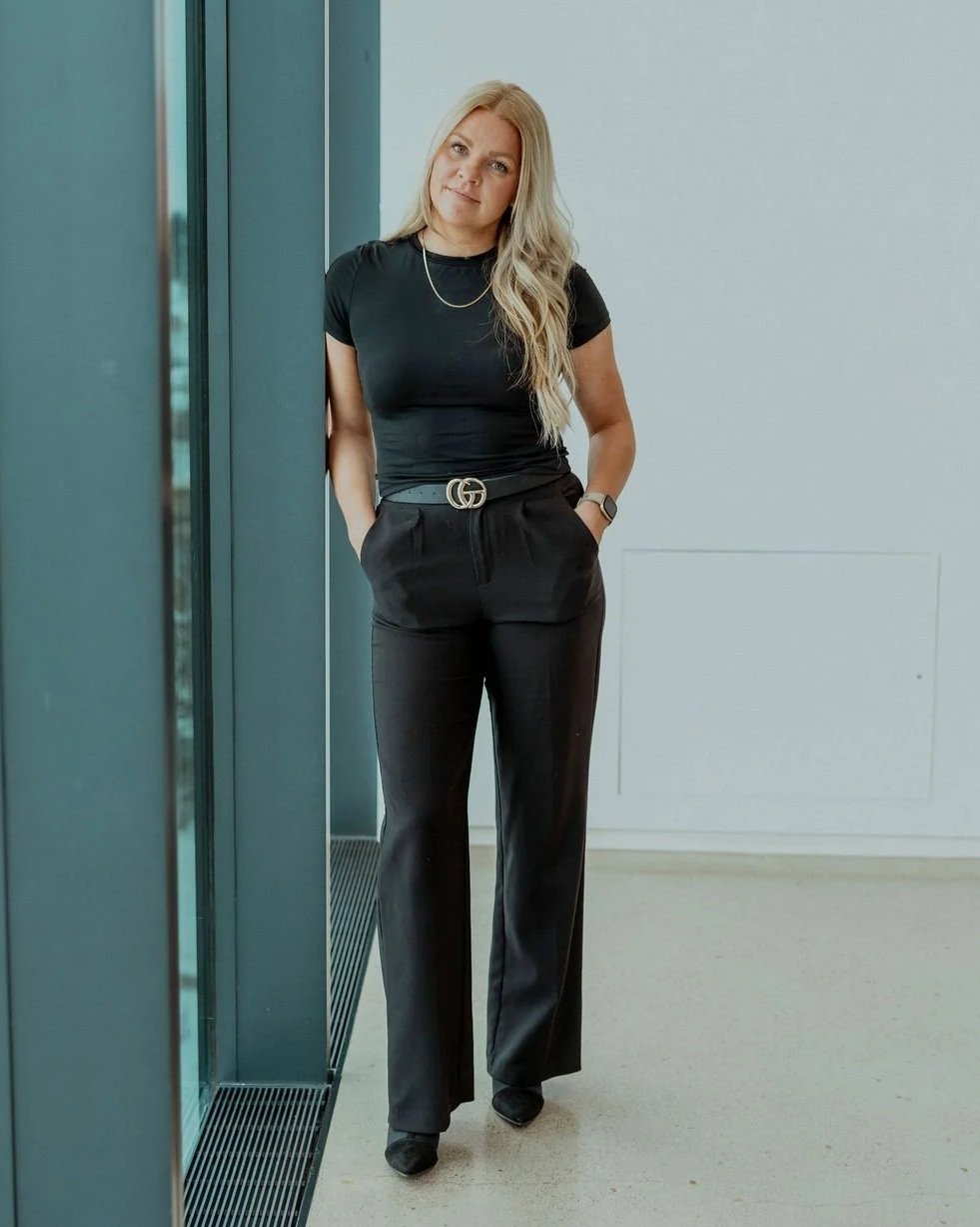 A woman with long blonde hair standing indoors near a blue wall and glass partition, wearing a black t-shirt, black high-waisted pants with a Gucci belt, and black shoes, with her hands in her pockets.
