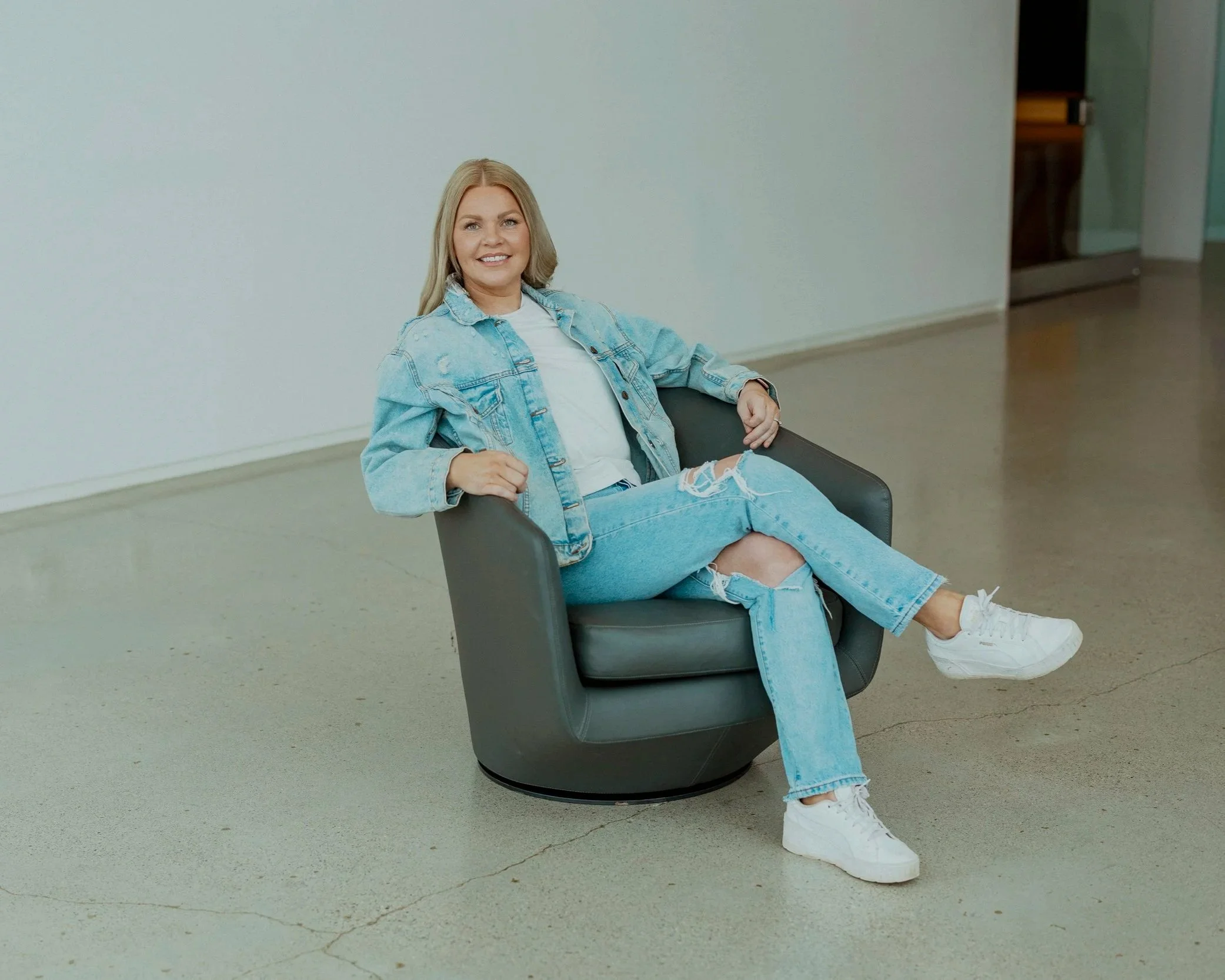 A woman with blonde hair wearing a denim jacket, white T-shirt, ripped jeans, and white sneakers sitting in a modern black swivel chair in a minimalistic room with a plain white wall and a polished concrete floor.