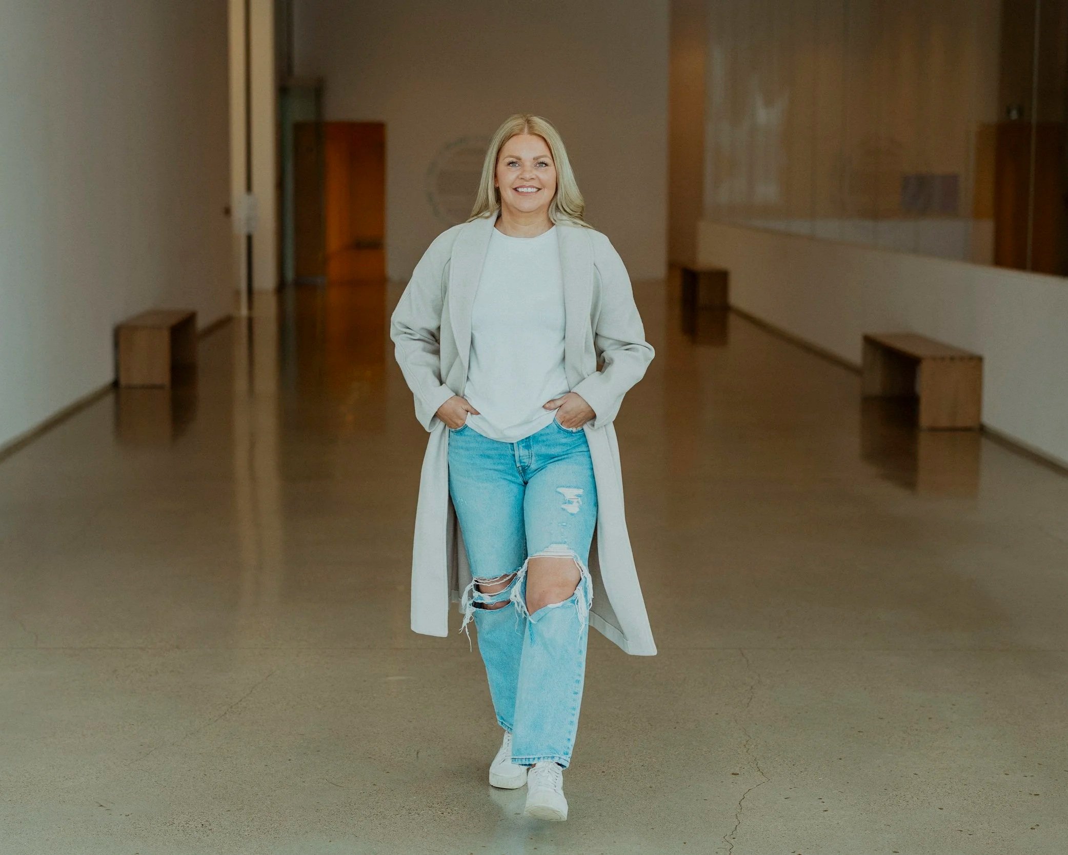 Woman with long blonde hair smiling, wearing a white sweater, light coat, ripped jeans, and white sneakers, walking in an empty indoor hallway with benches along the walls.