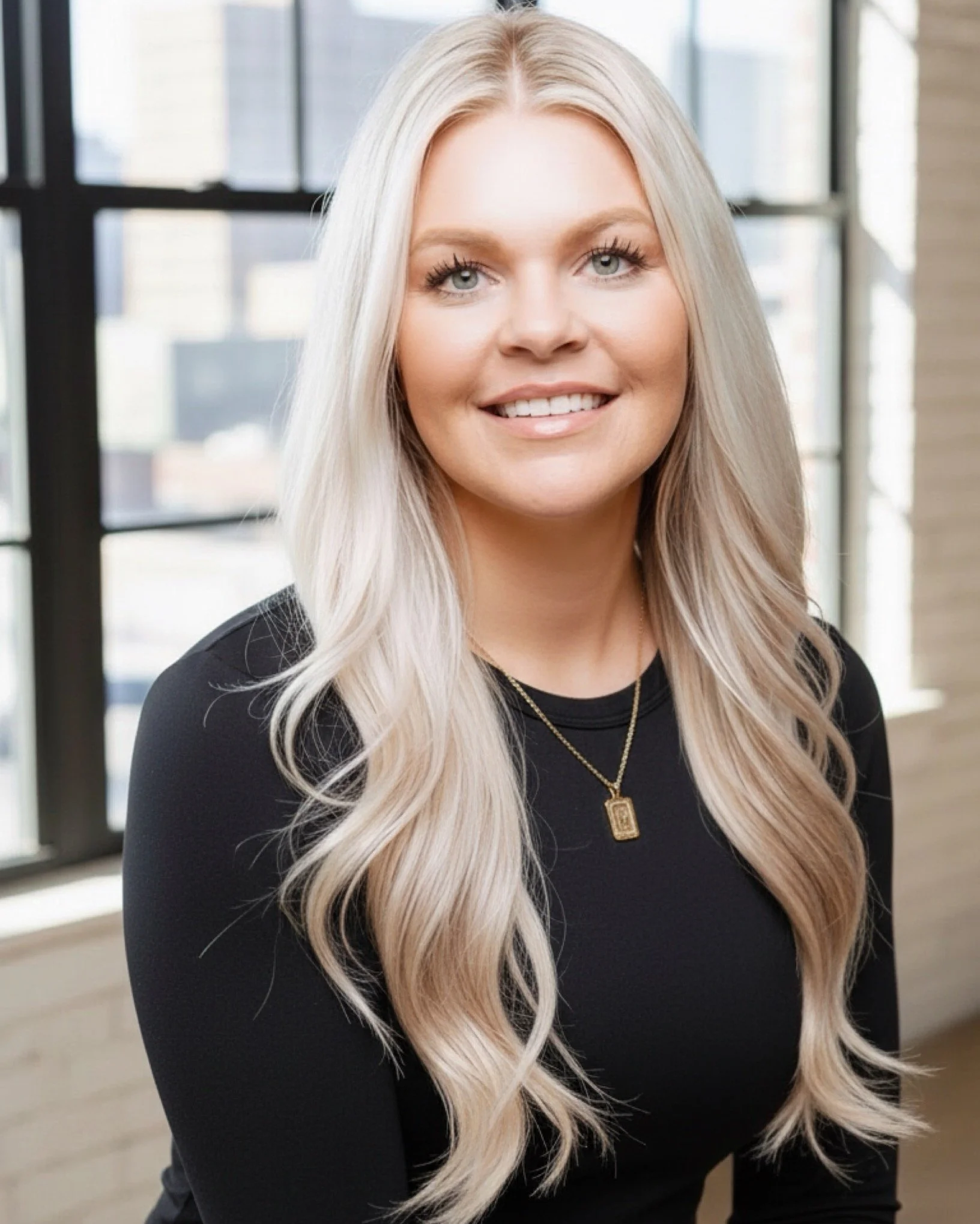 Portrait of a young woman with long blonde hair wearing a black top and a gold necklace, smiling, in an indoor setting with large windows and cityscape view in the background.