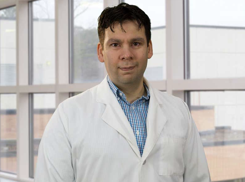 Portrait of a man wearing a white lab coat and blue checkered shirt standing indoors near large windows.