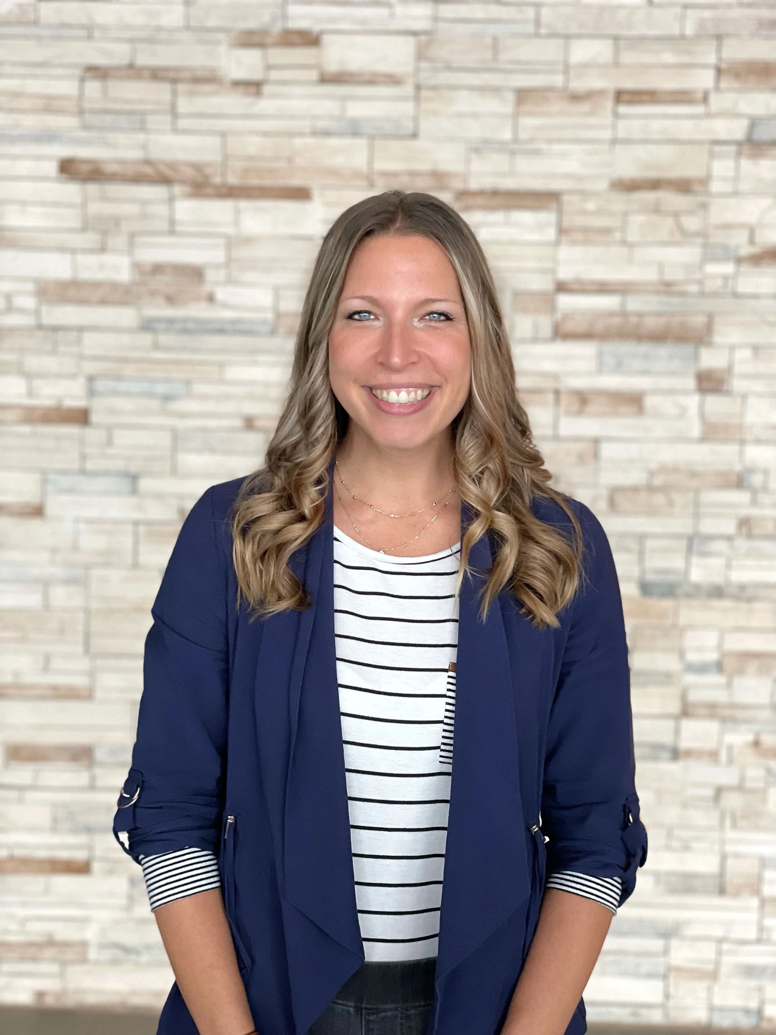 A woman with long, wavy blonde hair smiling, wearing a navy blazer over a white and black striped shirt, standing in front of a beige brick wall.