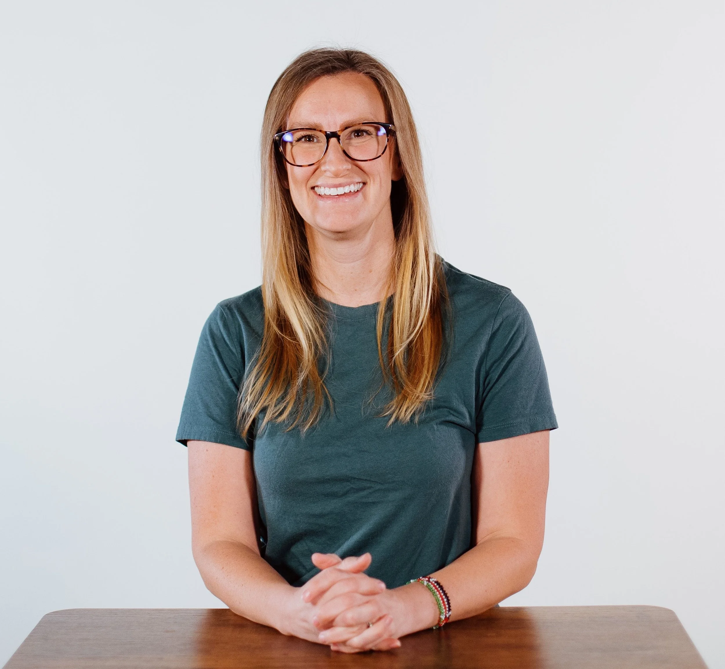 A woman with long blonde hair, wearing glasses and a dark green T-shirt, sitting at a wooden table, smiling at the camera, with a plain white background.