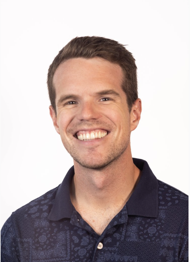 Smiling man with short brown hair, wearing a navy blue patterned polo shirt, looking at the camera against a plain white background.
