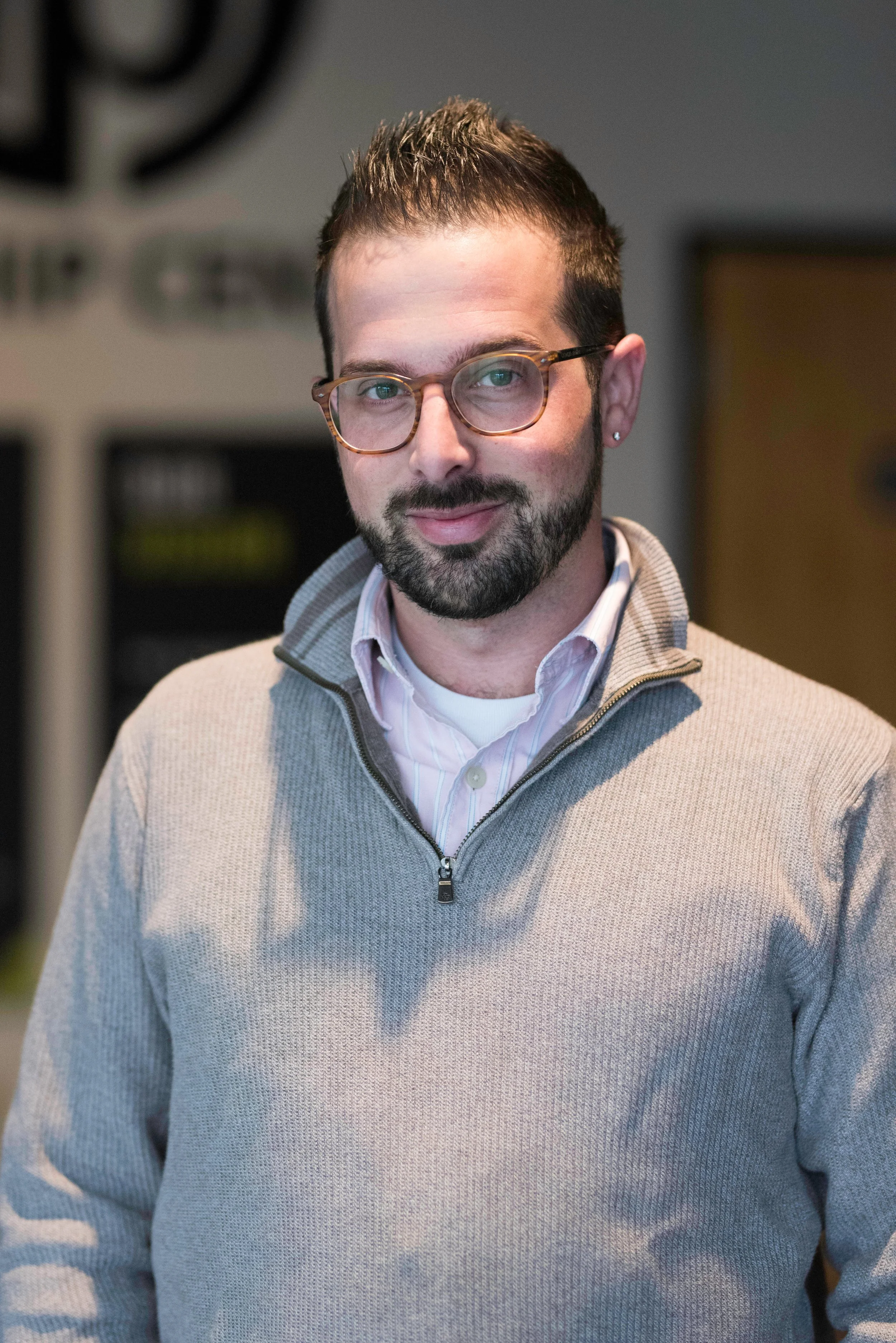 A man with glasses, beard, and short dark hair posing inside a building.