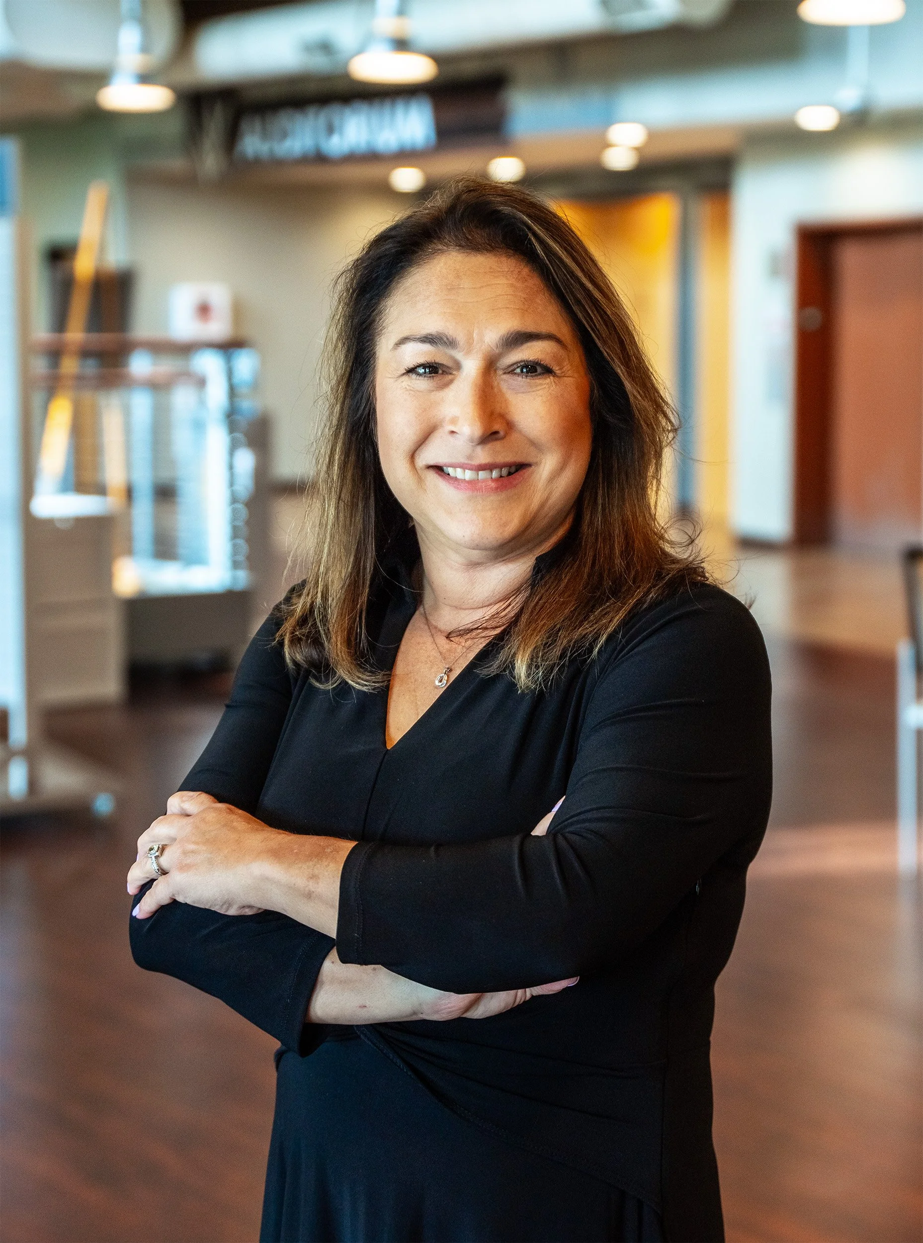 A woman in a black dress standing with arms crossed in a modern indoor setting with blurred background.
