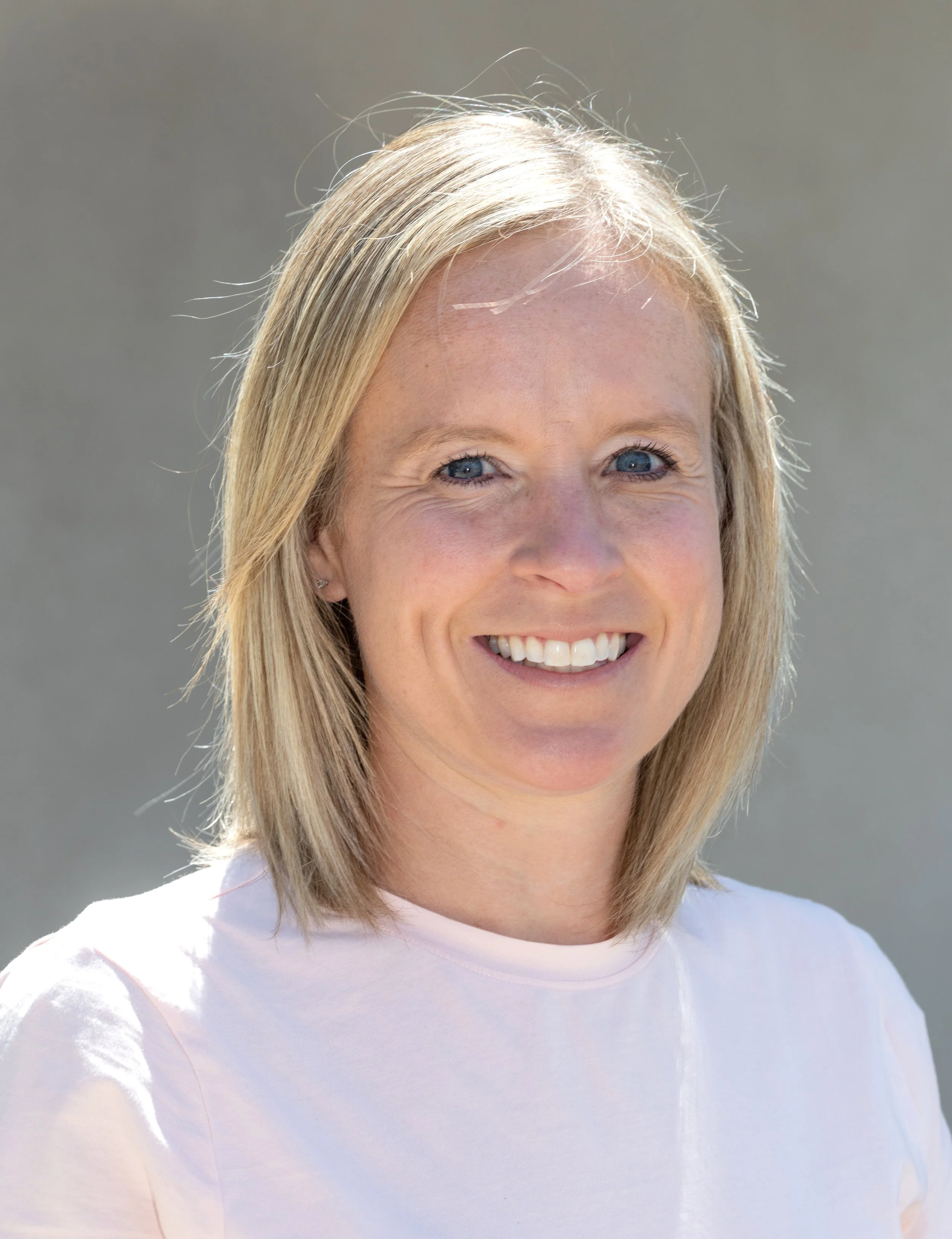 A woman with blonde hair and blue eyes smiles outdoors against a neutral background.